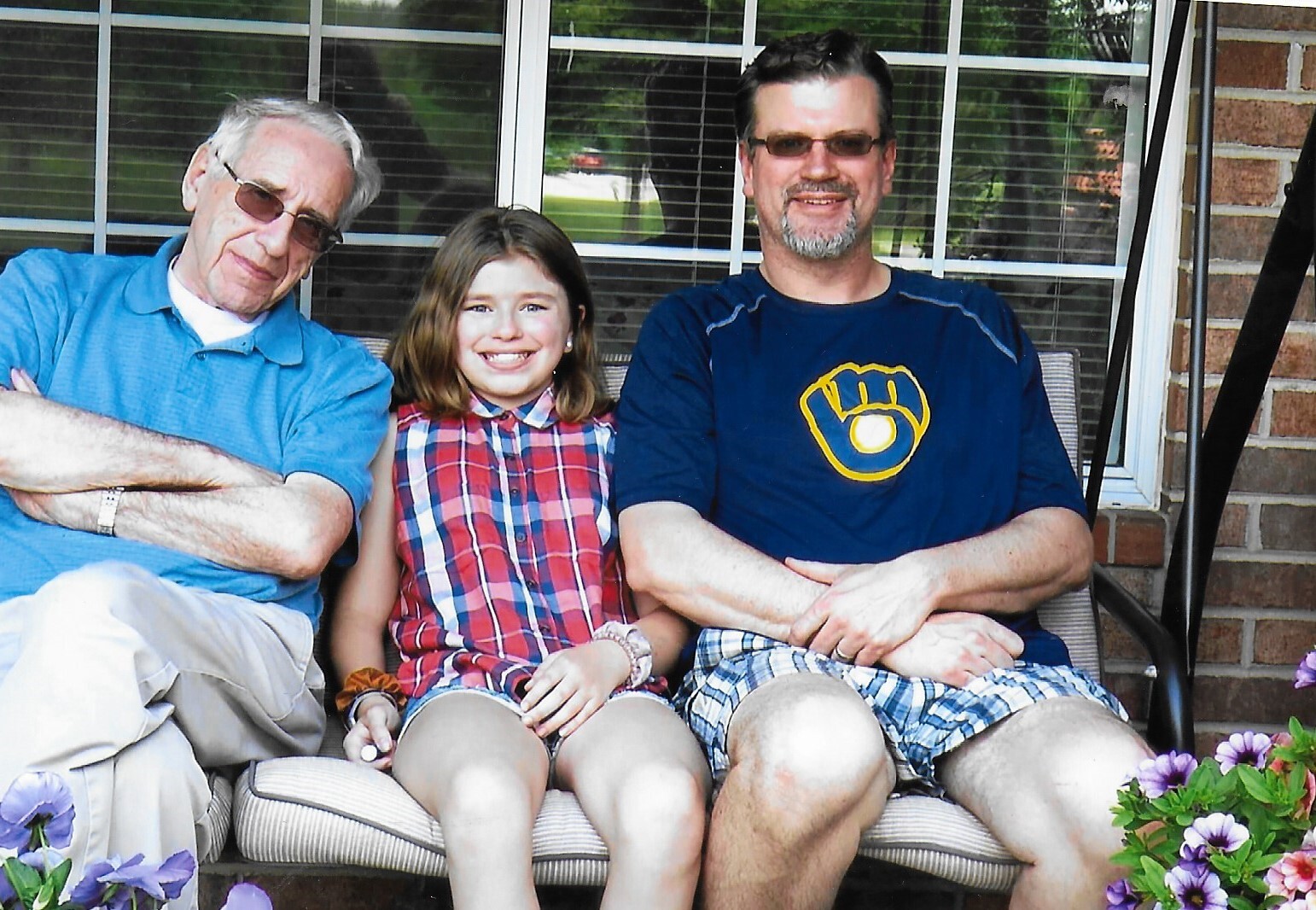Three people sit on a porch bench outside a brick house: grandfather in blue polo, smiling girl in plaid, father in dark shirt with logo and shorts, all smiling at the camera.
