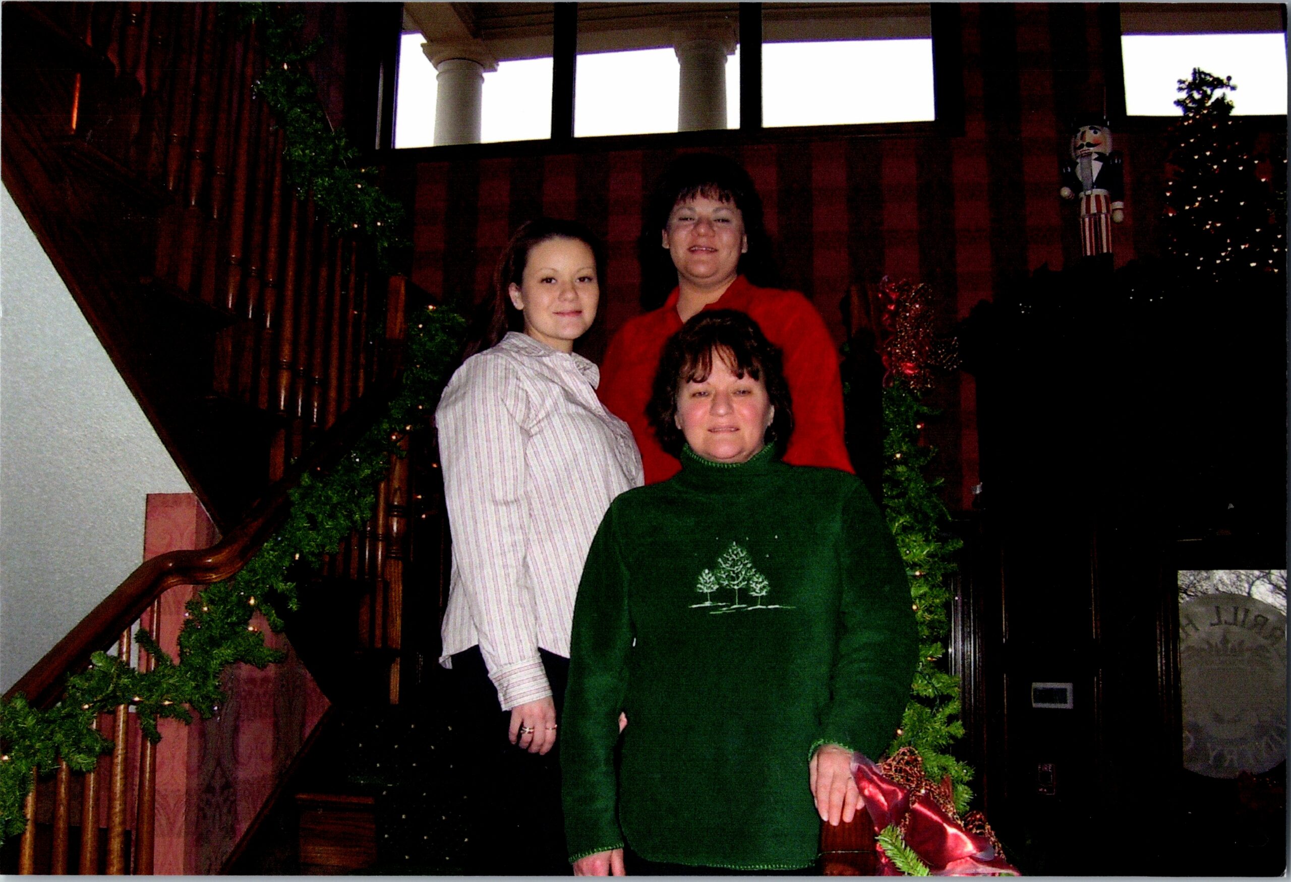 Three women posing on a decorated indoor staircase with garlands; one in a green Christmas sweater in front, two behind (striped shirt and red top).