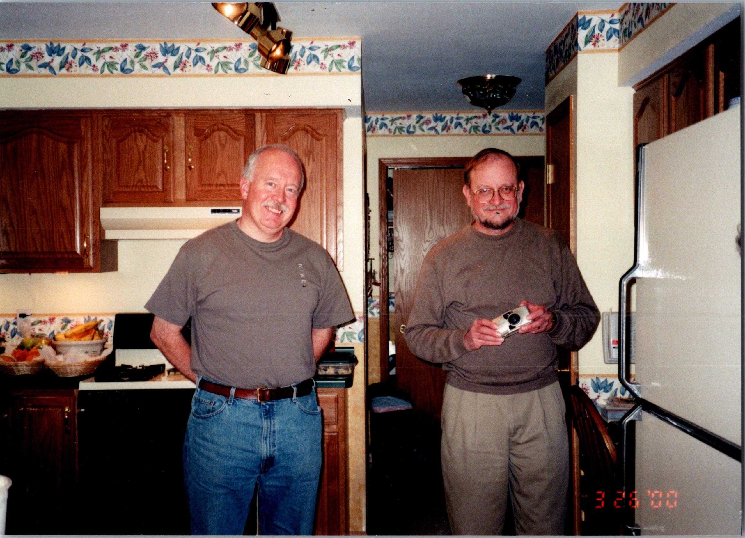Two men stand in a kitchen smiling at the camera, with wooden cabinets and a fruit bowl in the background.