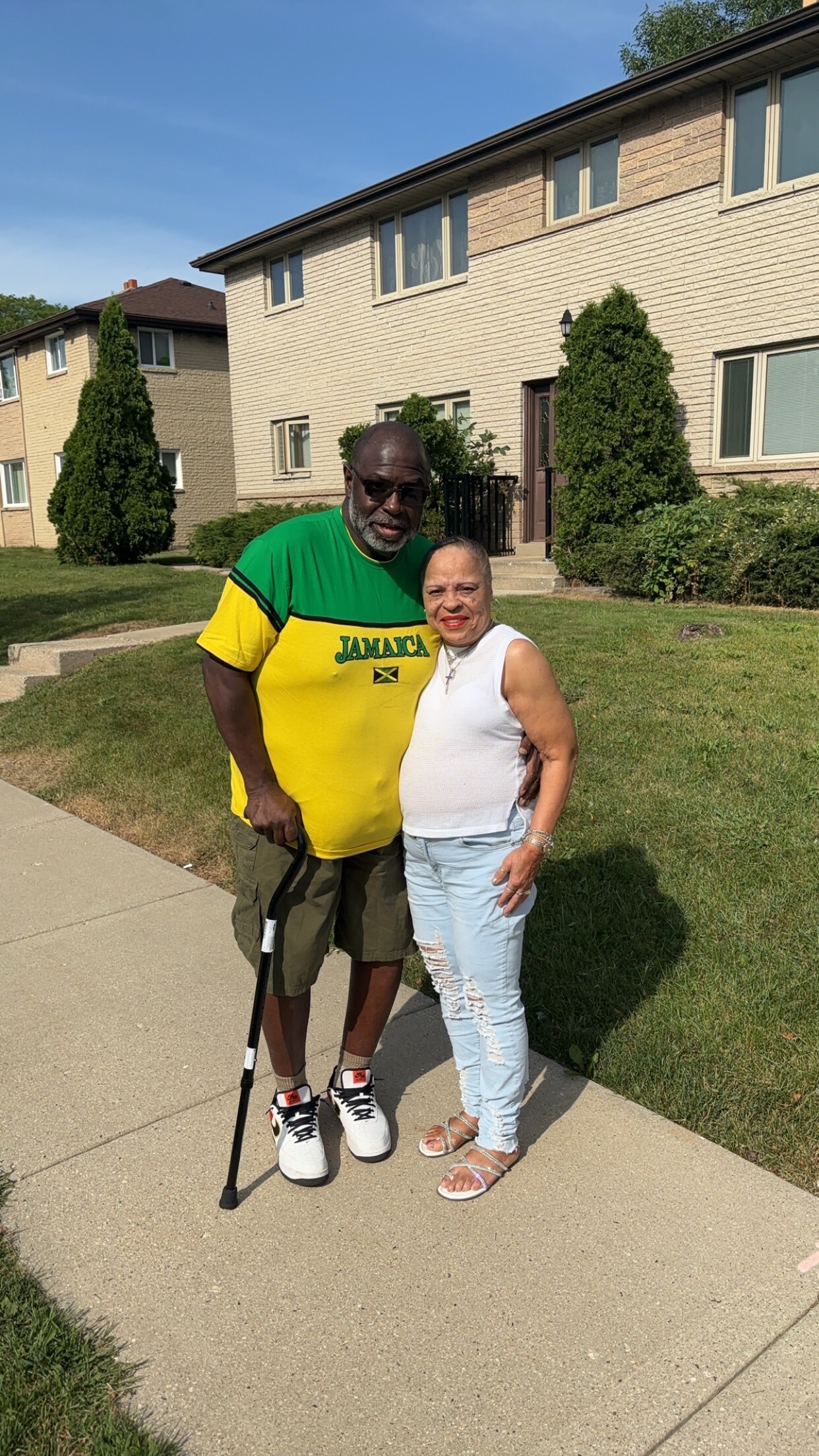 Two adults pose together on a sidewalk in front of a beige apartment building; the man wears a Jamaica shirt and holds a cane, the woman in a white sleeveless top smiles beside him.