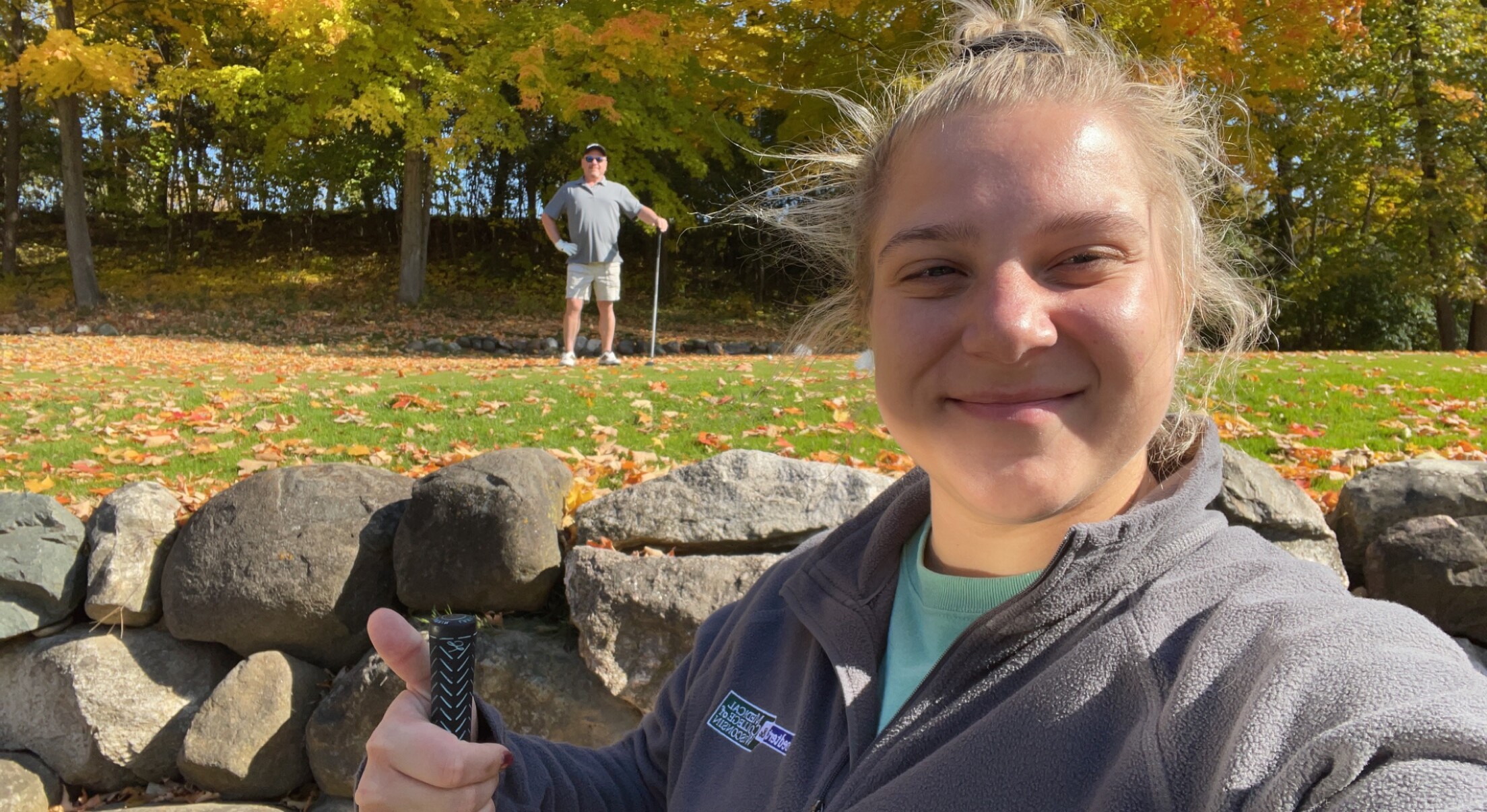 Close-up selfie of a smiling woman in the foreground with autumn leaves and a man standing with a golf club in the background.