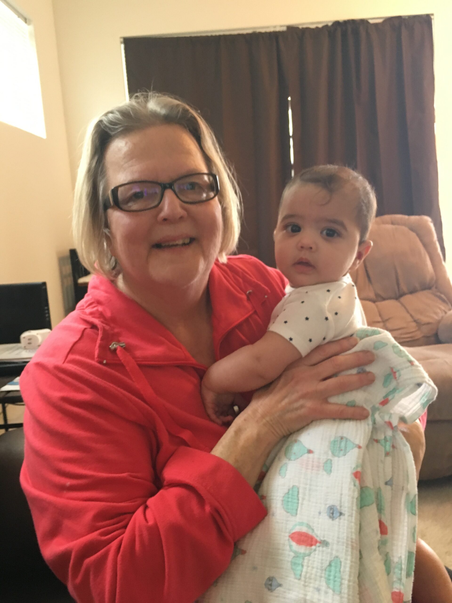 Older woman wearing glasses and a red shirt holding a baby wrapped in a pastel blanket indoors, both looking at the camera.