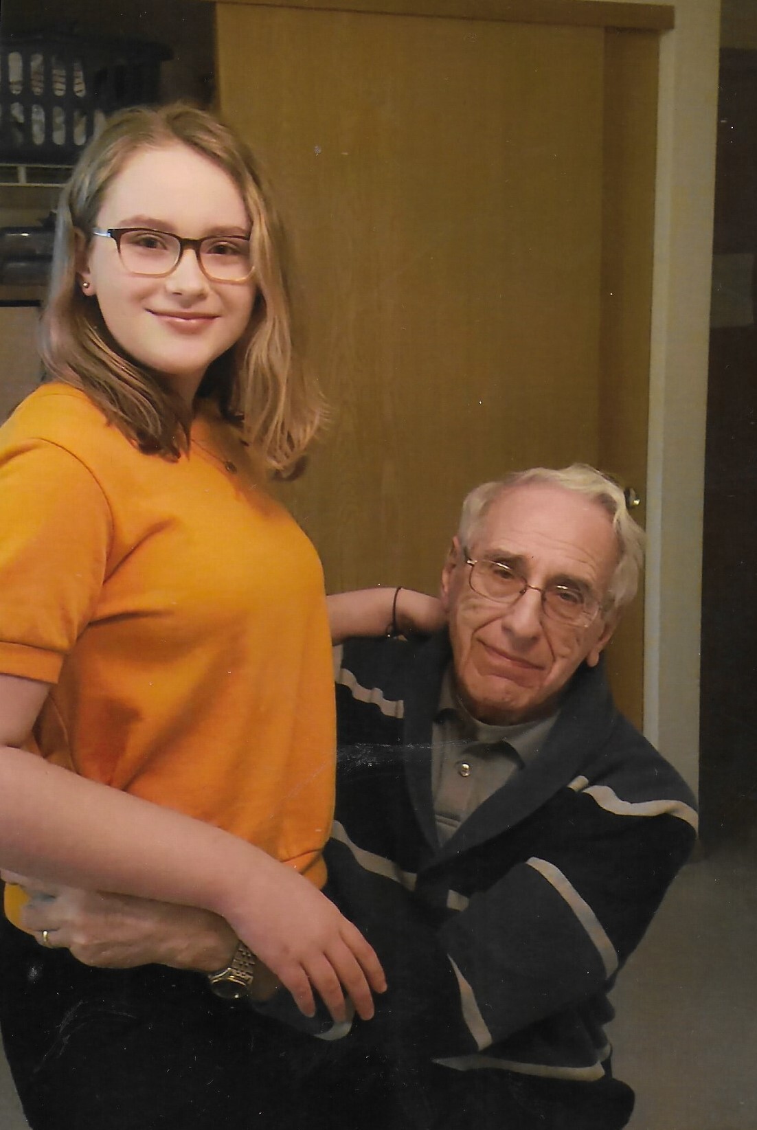 A young woman in an orange shirt stands beside an older man who sits and rests his arm around her, smiling softly indoors.