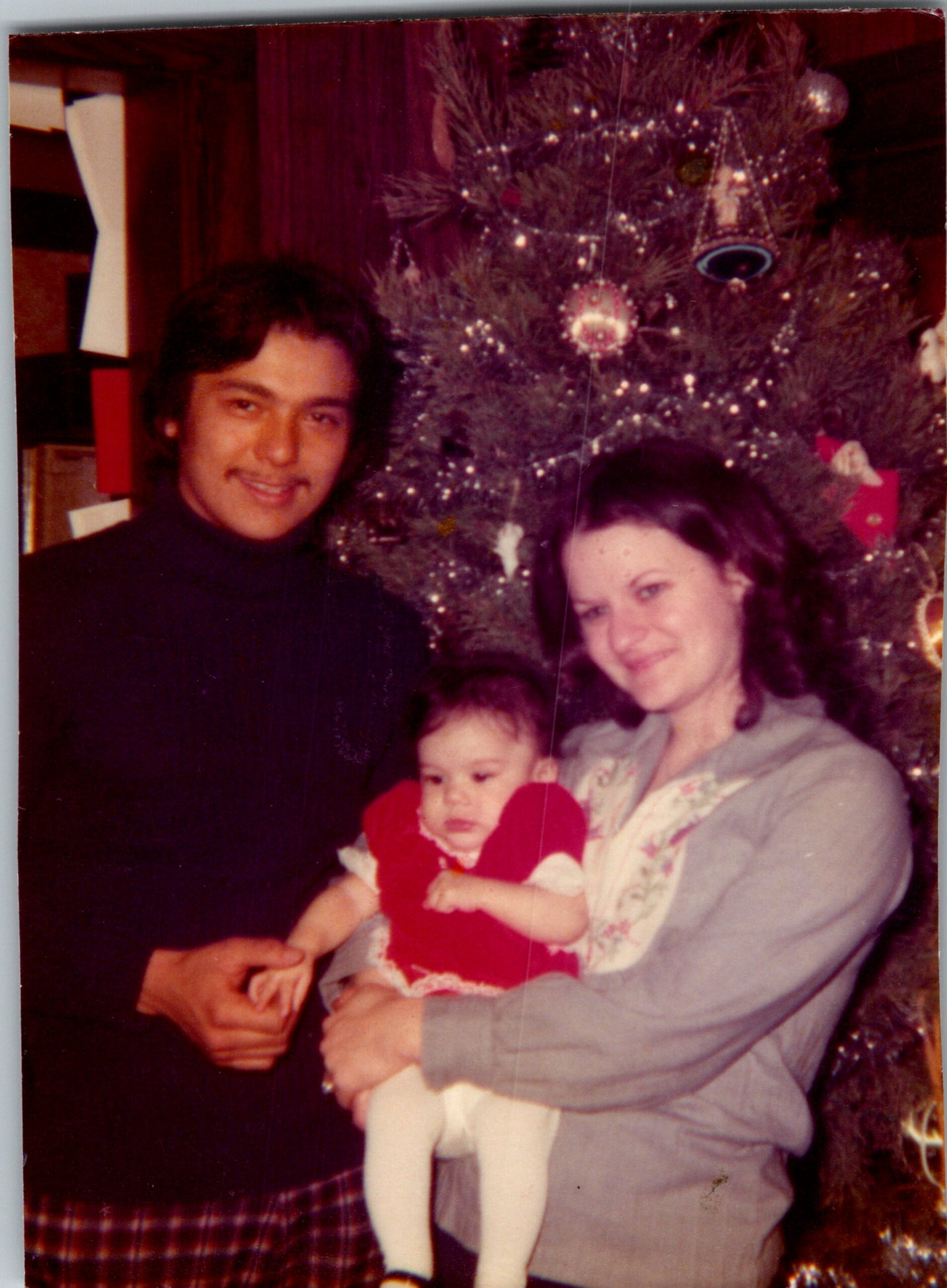 Man and woman hold a baby in front of a decorated Christmas tree indoors.