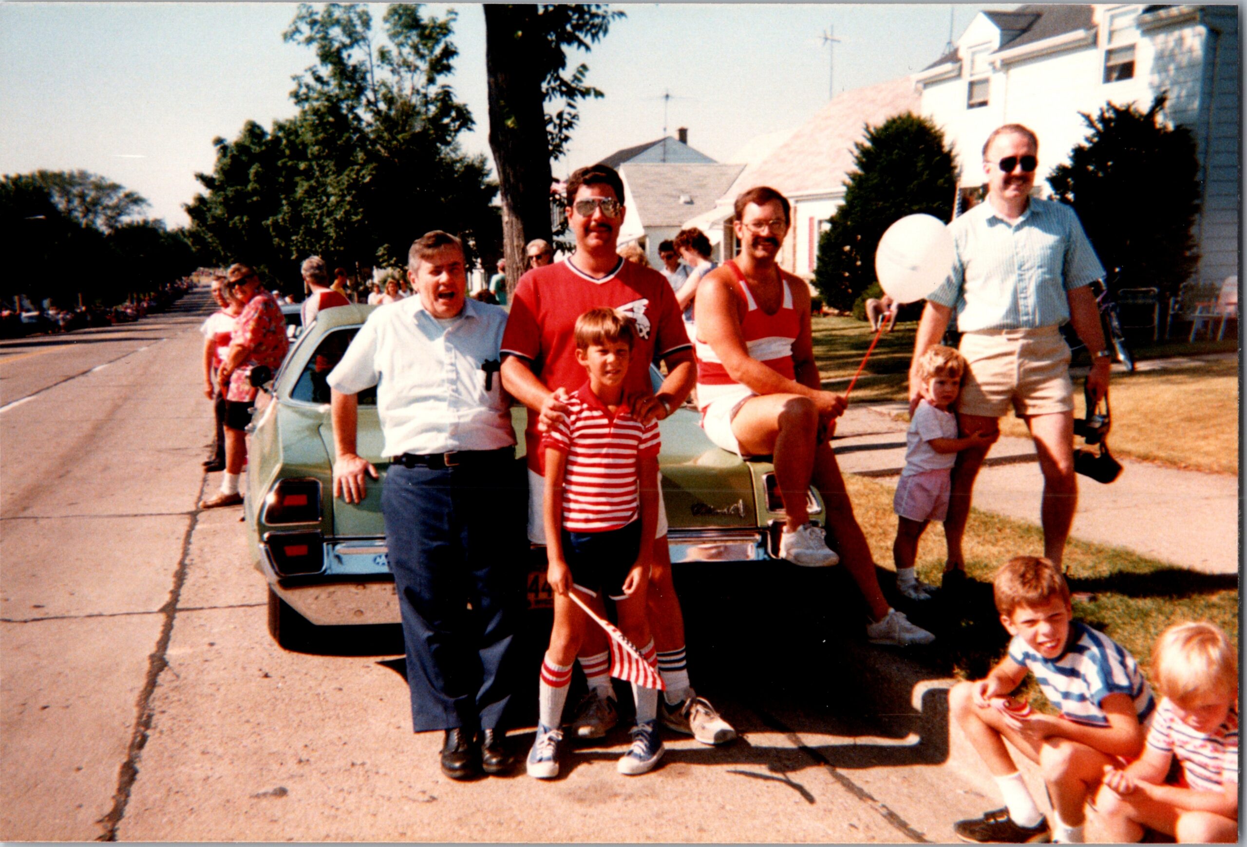 Group of people posing in front of a vintage car on a sunny street, kids in front and adults smiling at a parade or gathering