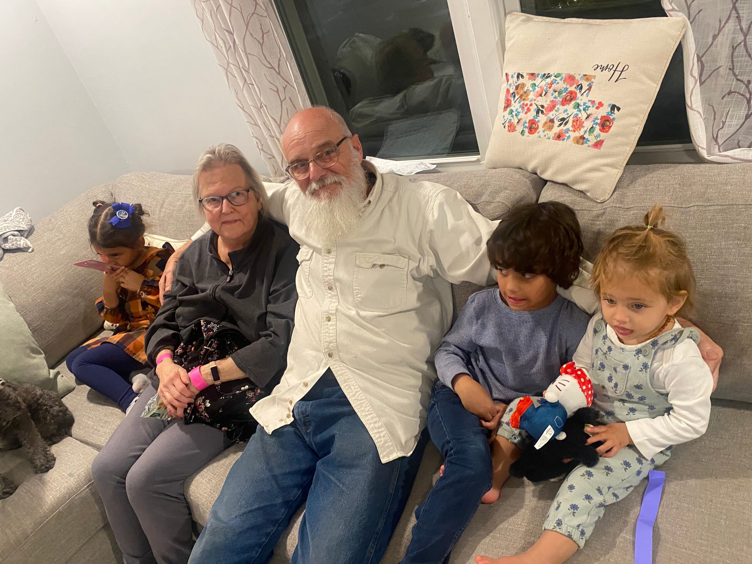 Grandfather with glasses and white beard sits beside a woman on a gray sofa, flanked by two children in a living room.