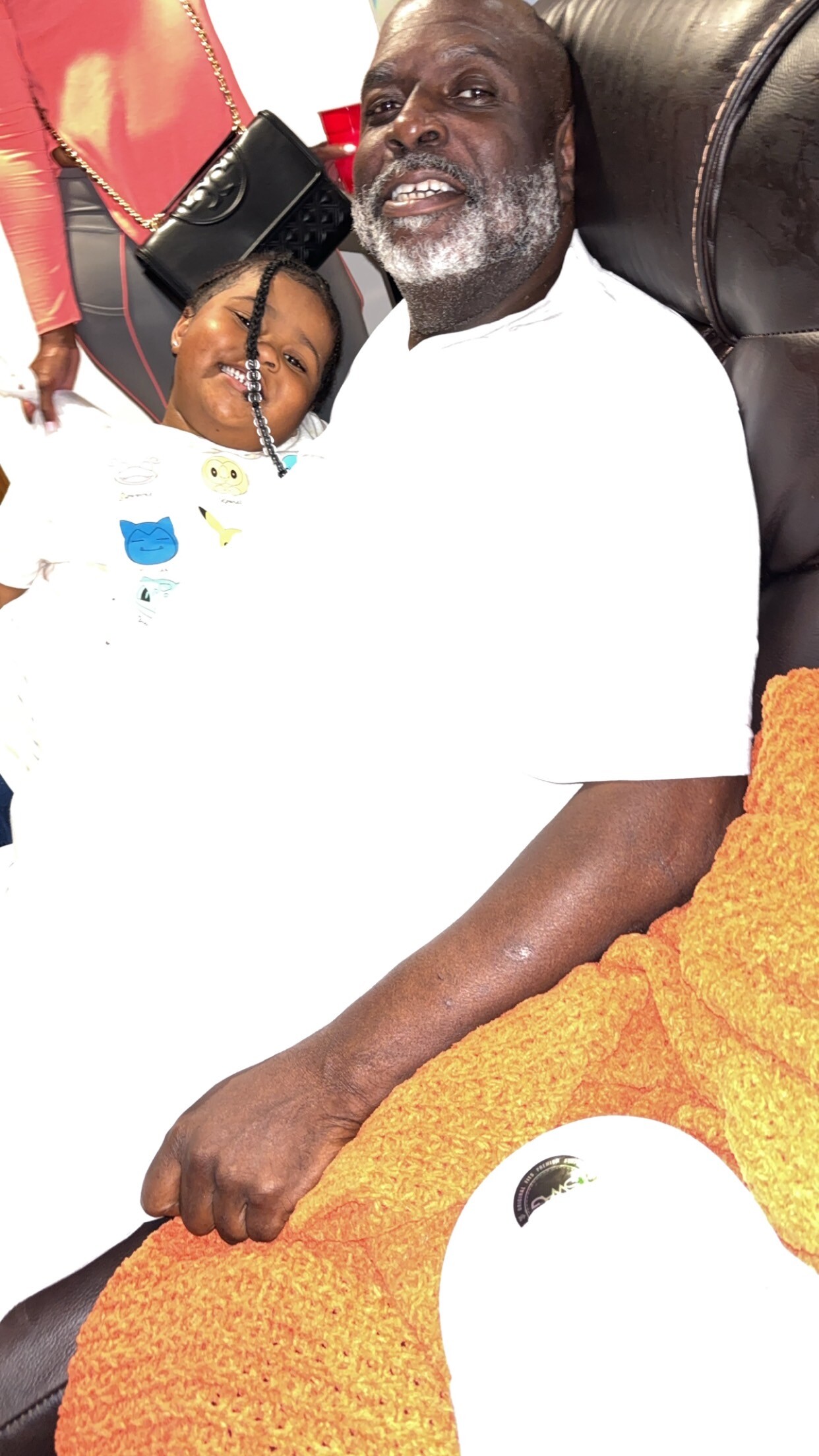 Older man with a gray beard wearing a white shirt sits on a brown leather chair, a child leaning on him, and an orange towel in the foreground.