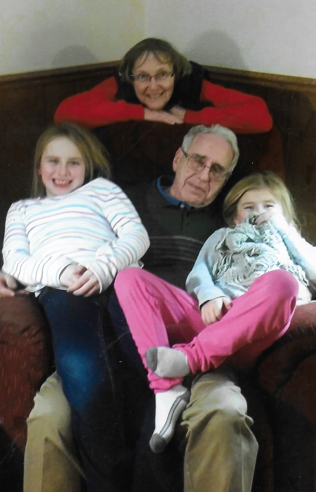 An older man sits on a brown sofa with two smiling young girls, a woman in red leaning over the back of the sofa behind them.