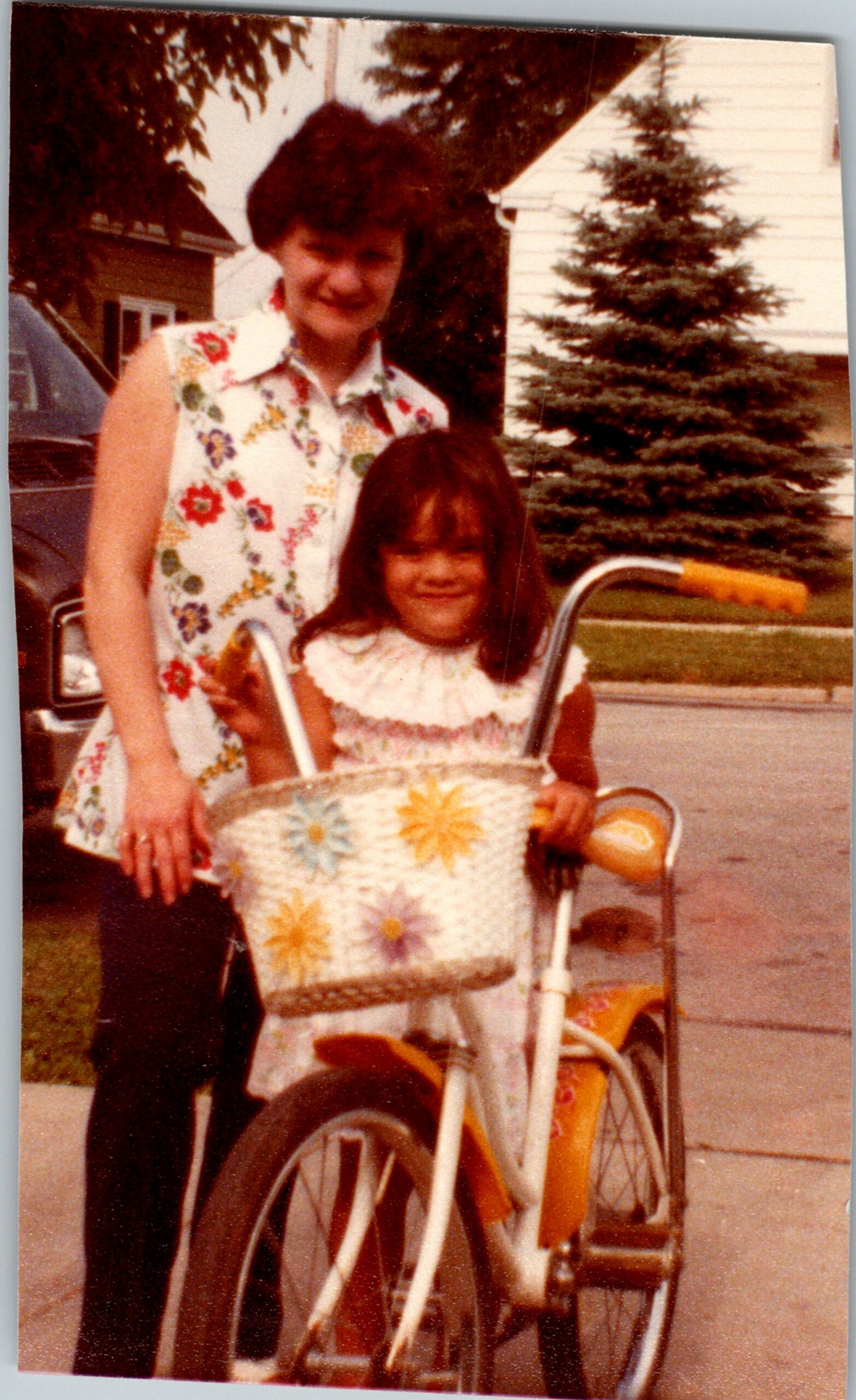 Smiling woman in a floral sleeveless dress stands beside a young girl outdoors near a bicycle with a wicker basket in a suburban yard.