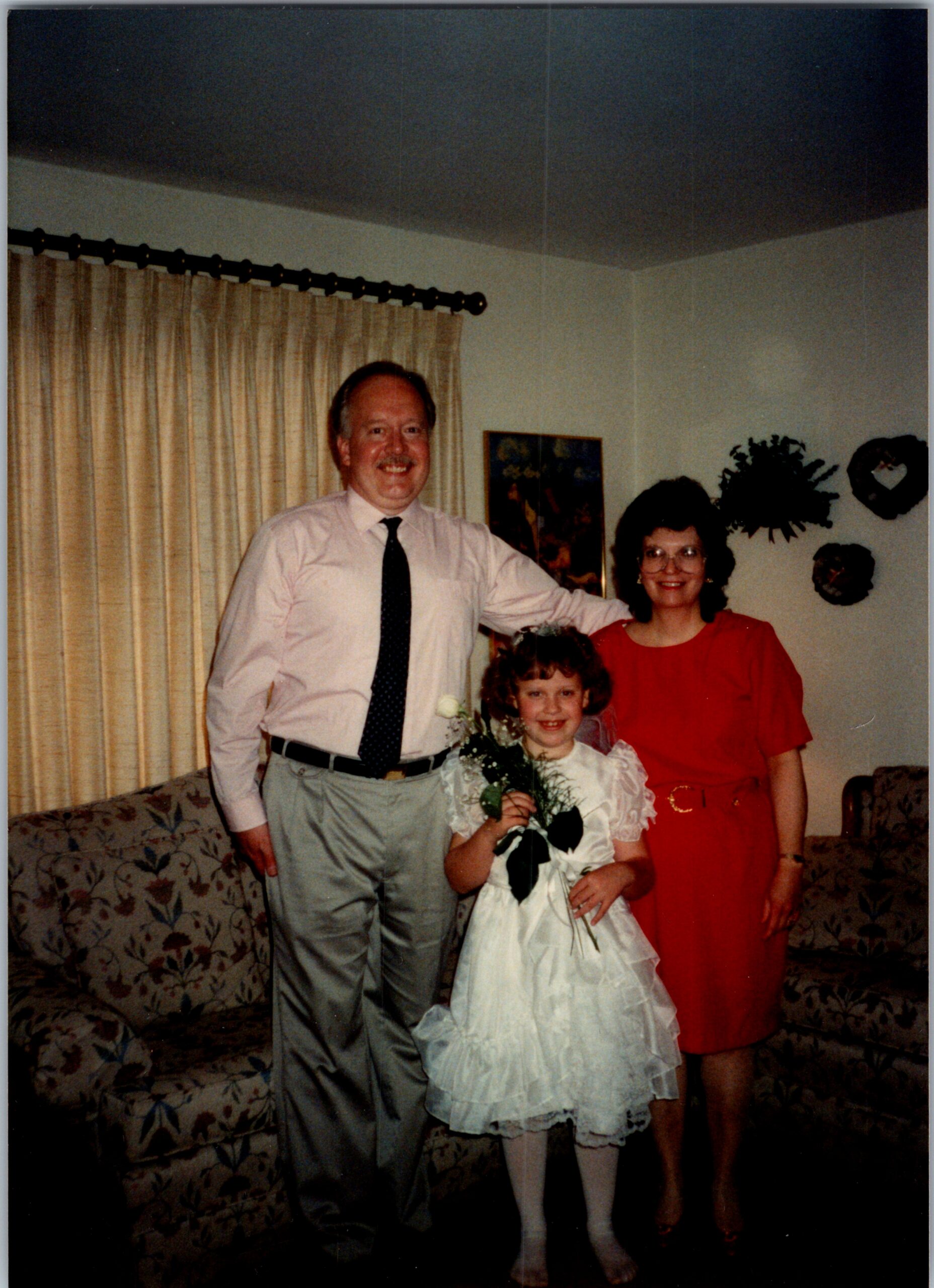 Family posing for a celebratory photo indoors: man in a white shirt and tie, woman in a red dress, and a girl in a white dress holding a bouquet, standing in front of a patterned sofa and beige curtains.