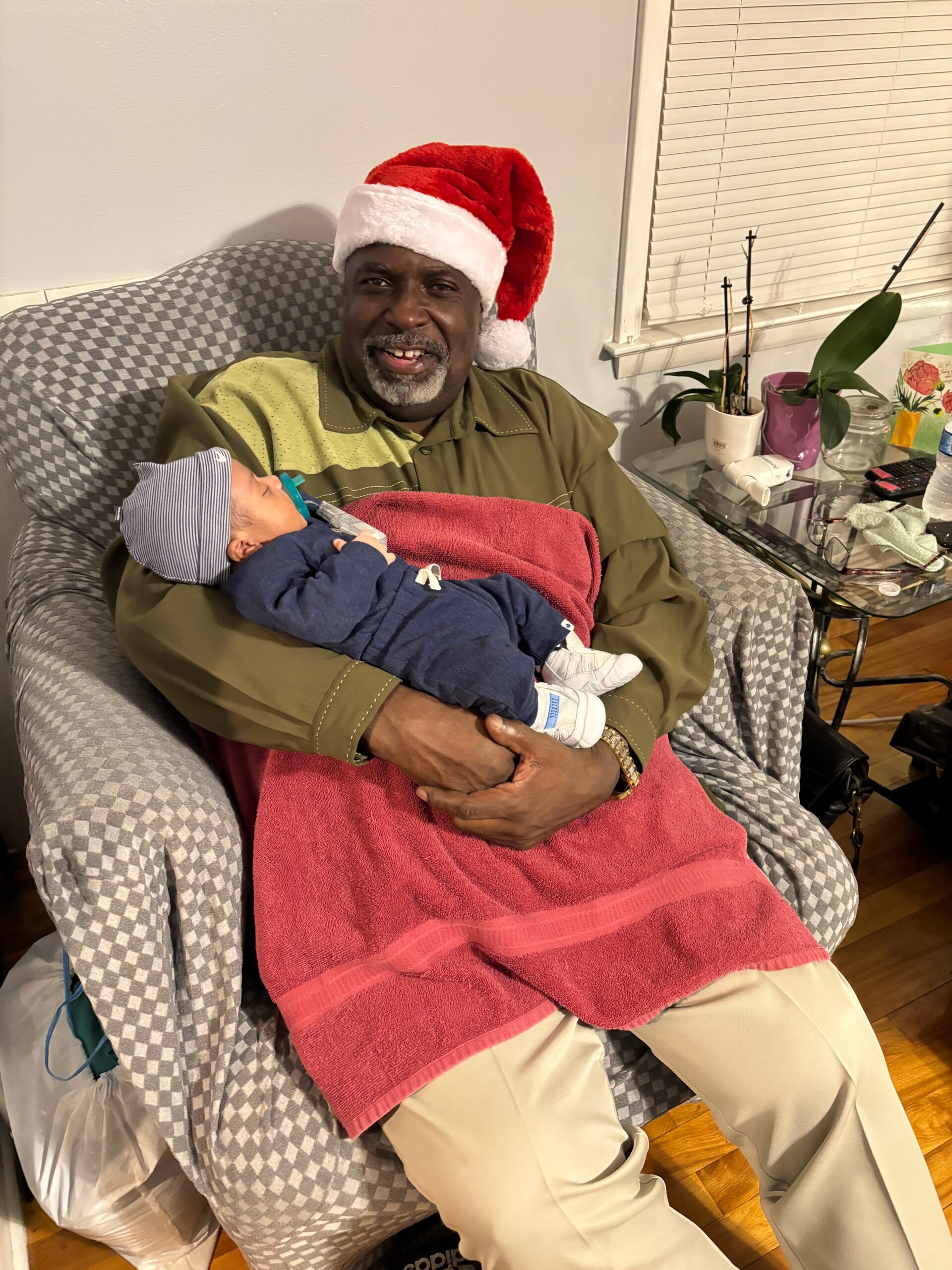 Man in a Santa hat smiles while holding a baby wrapped in a red blanket on his lap in a living room setup.