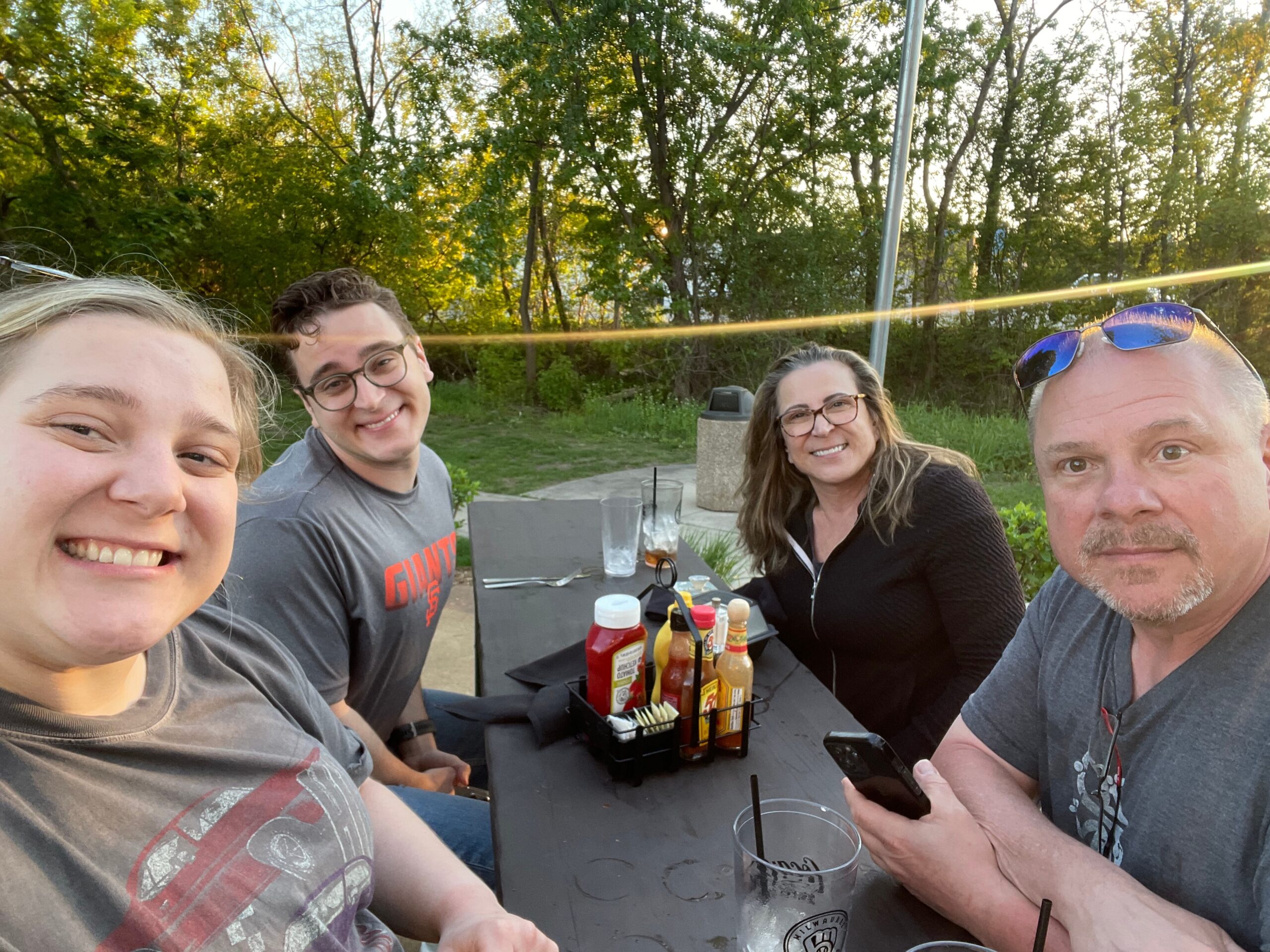 Four adults sit around an outdoor picnic table, smiling at the camera with condiments and drinks on the table, trees in the background.