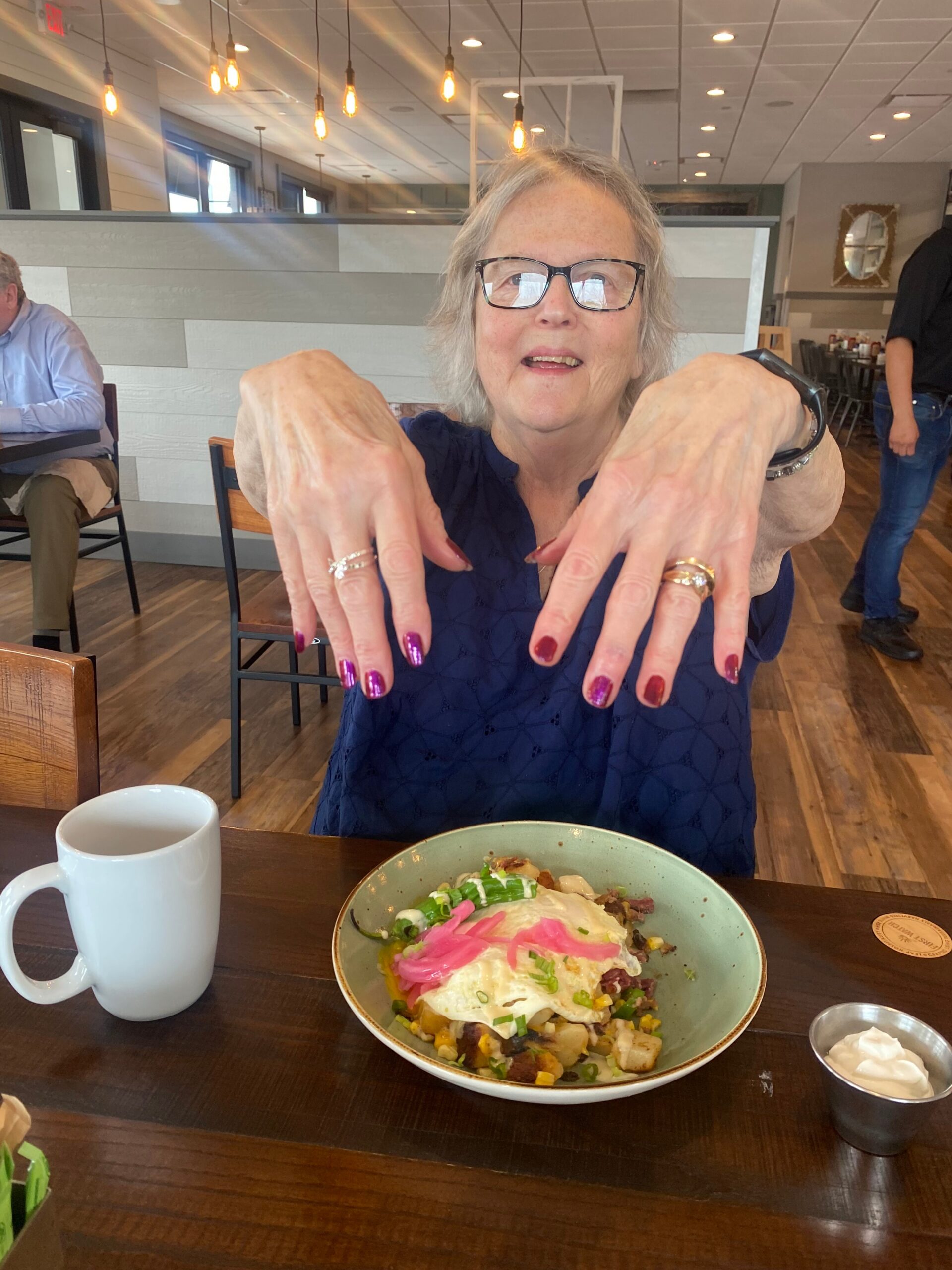 Smiling elderly woman with glasses shows her painted nails at a restaurant, hands raised toward the camera with a plated dish in front.
