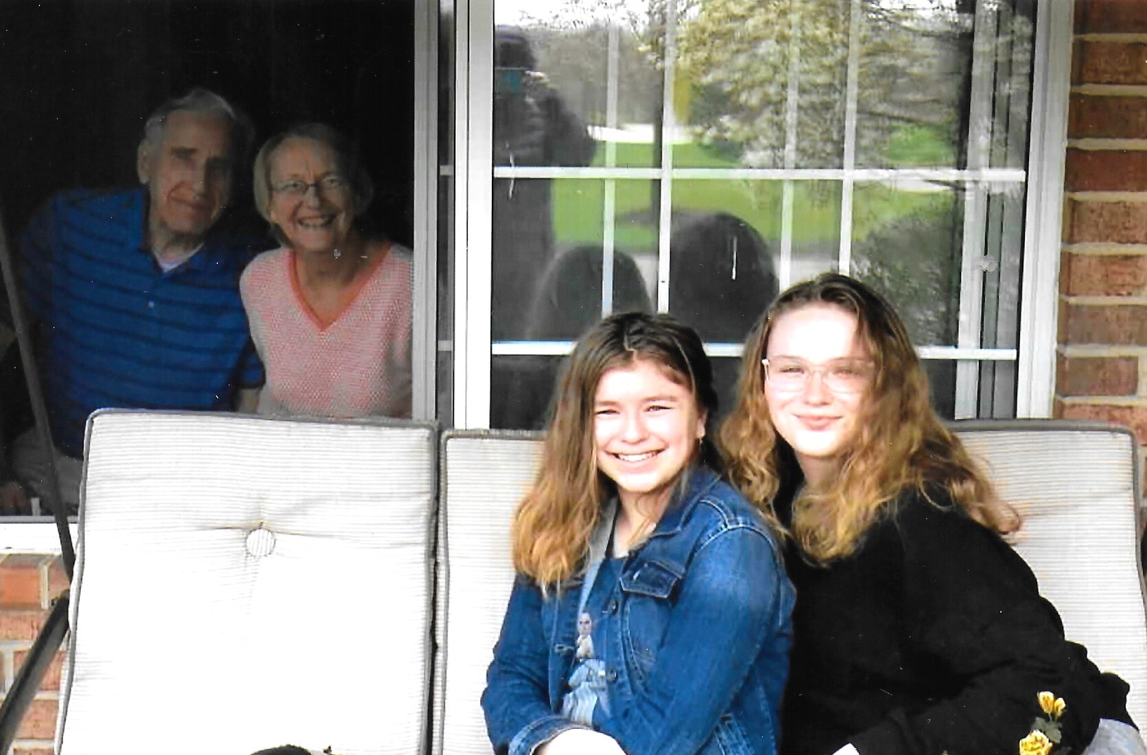 Two young women pose on a porch chair while an older couple looks out from a window behind them.