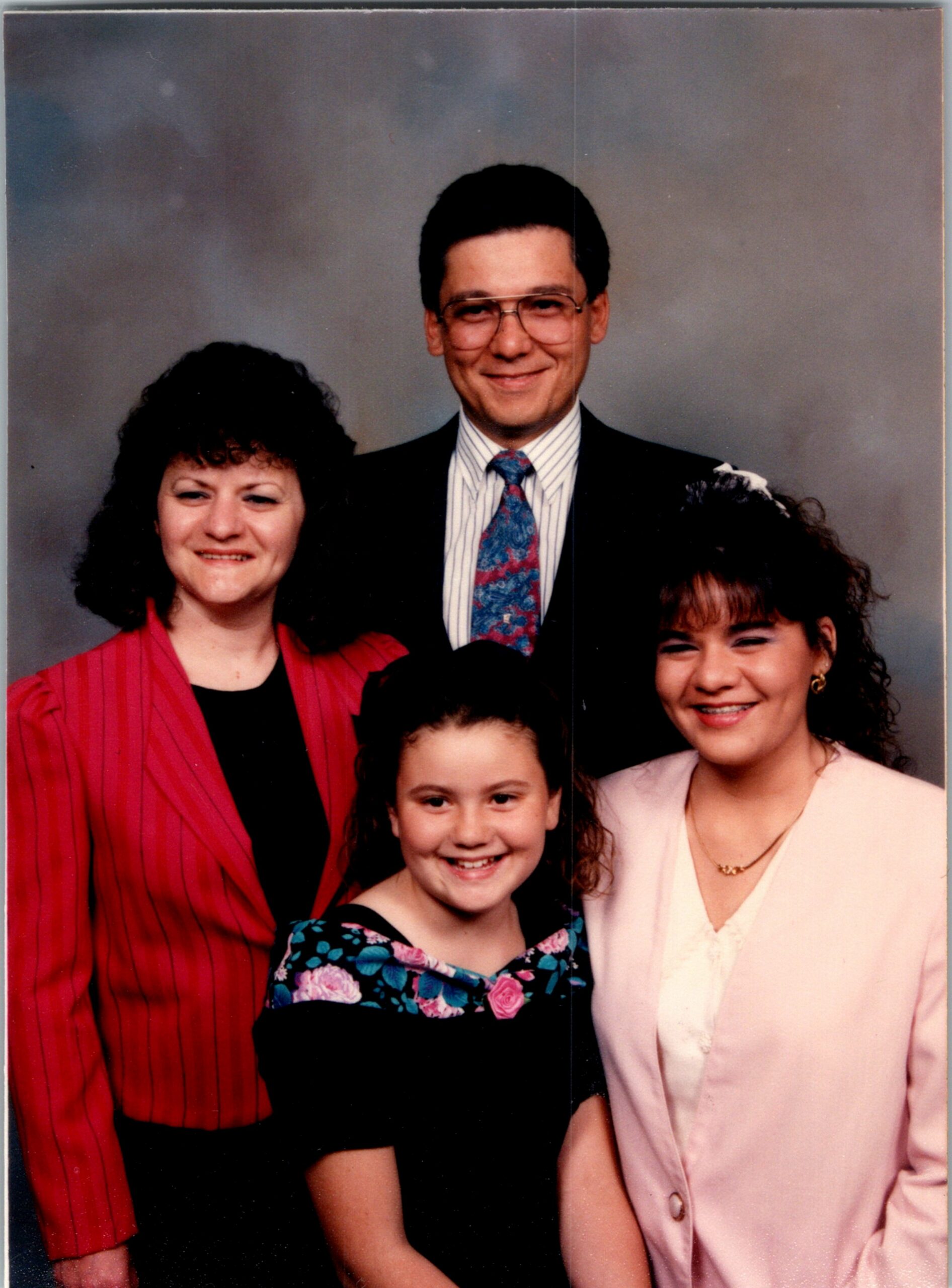 Family portrait of four smiling people in formal clothes against a neutral studio backdrop, including two women, a man, and a girl in dresses.