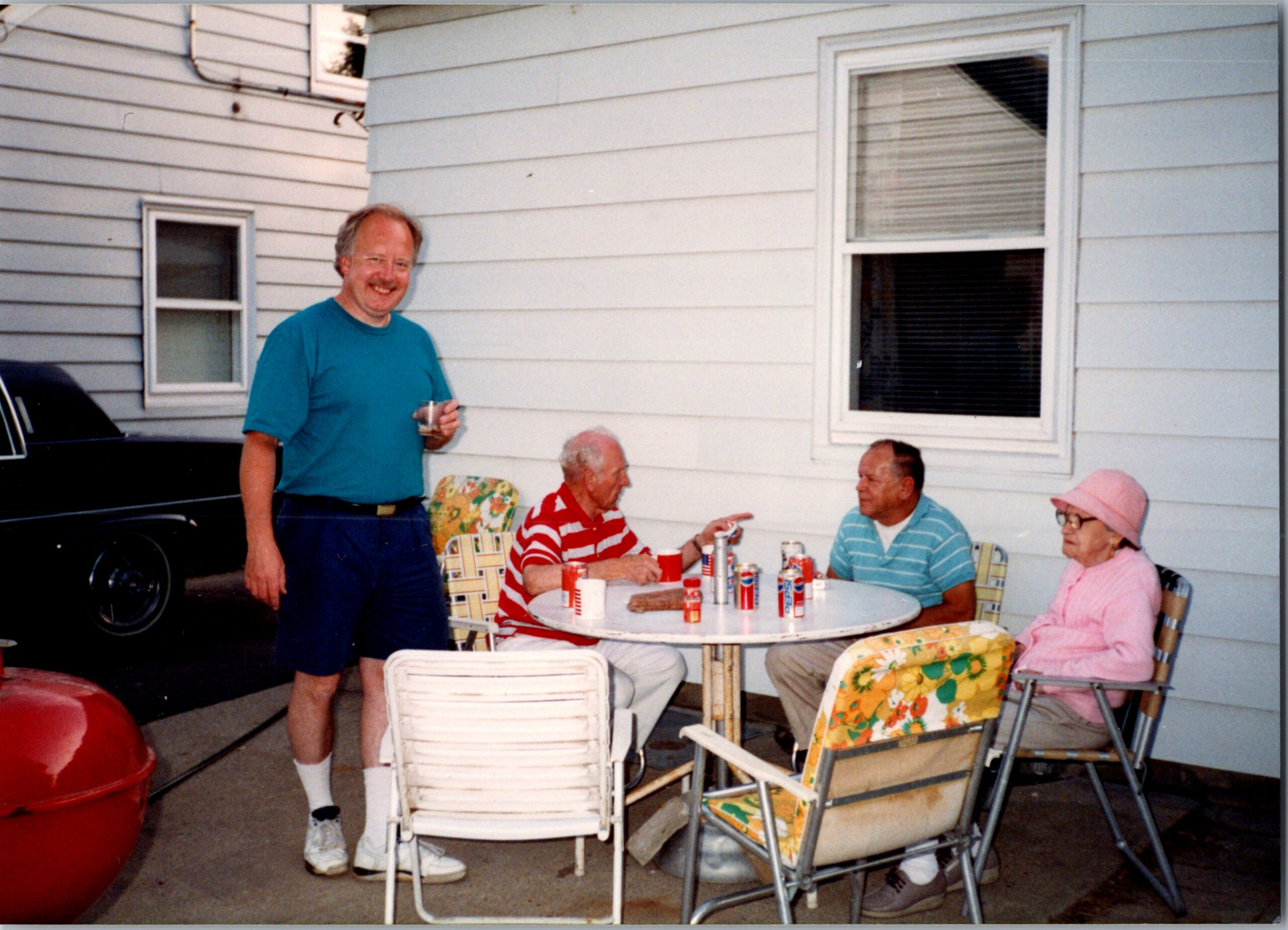 Smiling man in a teal shirt standing beside a round table of seniors sitting outdoors on a sunny patio.