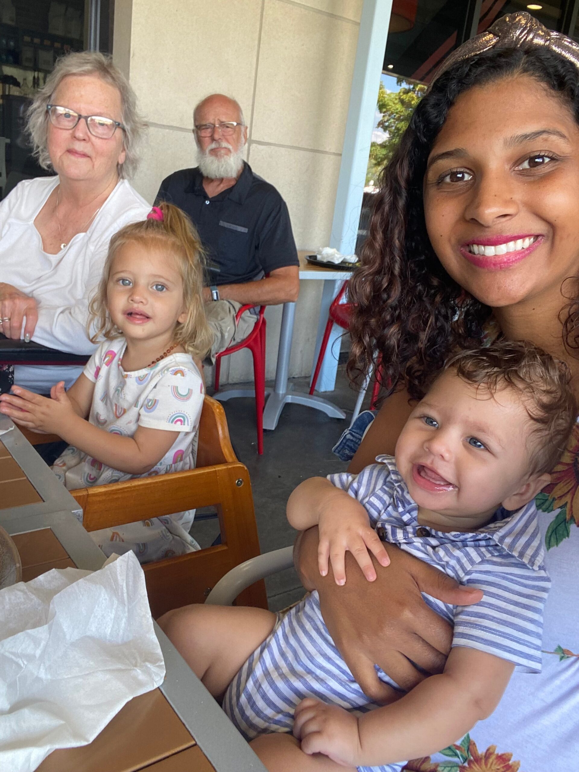 Young woman takes a selfie with a smiling toddler and a little girl at a table outdoors, while an elderly couple sits behind them.