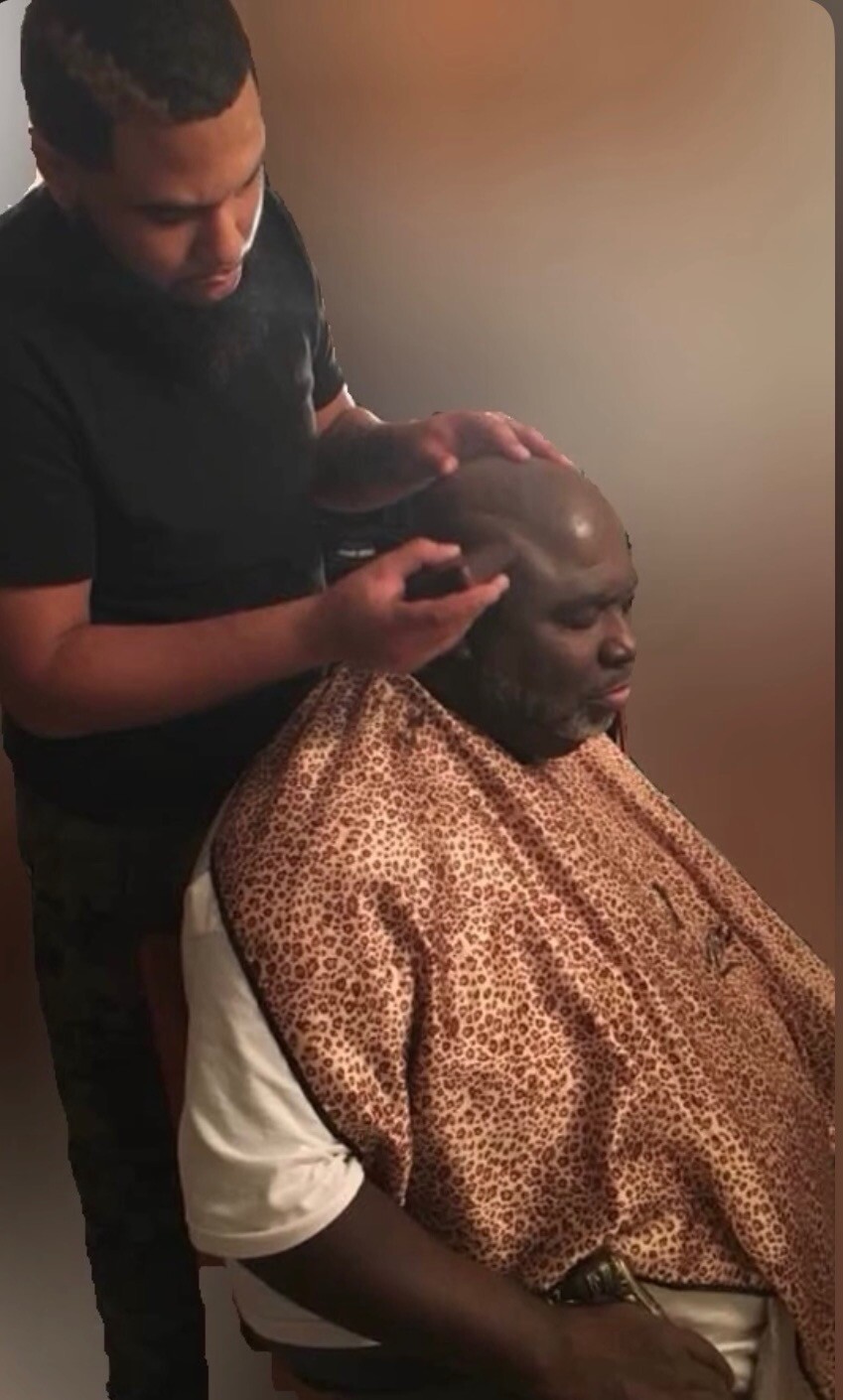 Barber trimming a bald man’s scalp while he sits wearing a leopard-print barber cape.