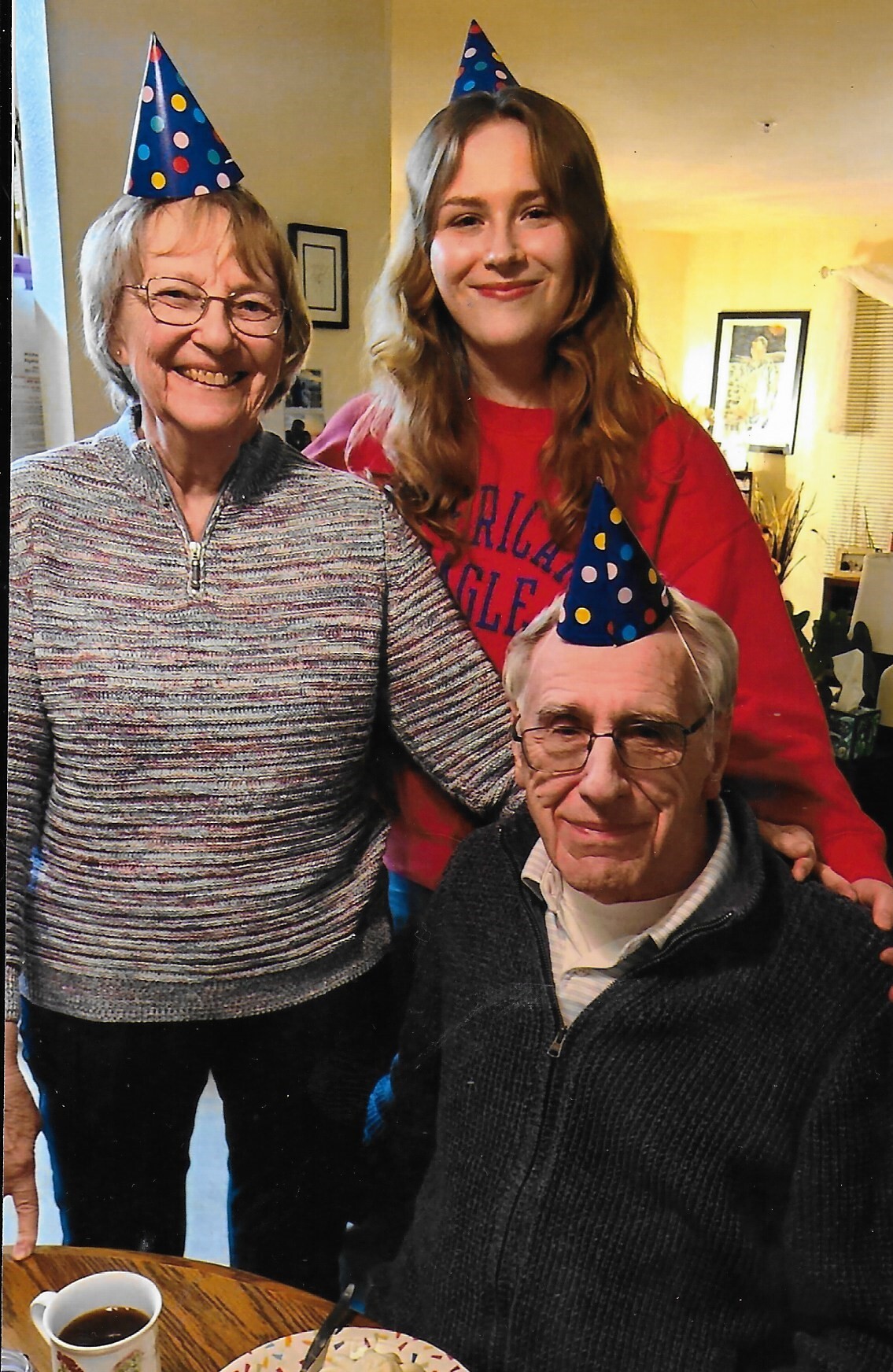 Three people posing for a birthday photo, wearing colorful party hats and smiling indoors.