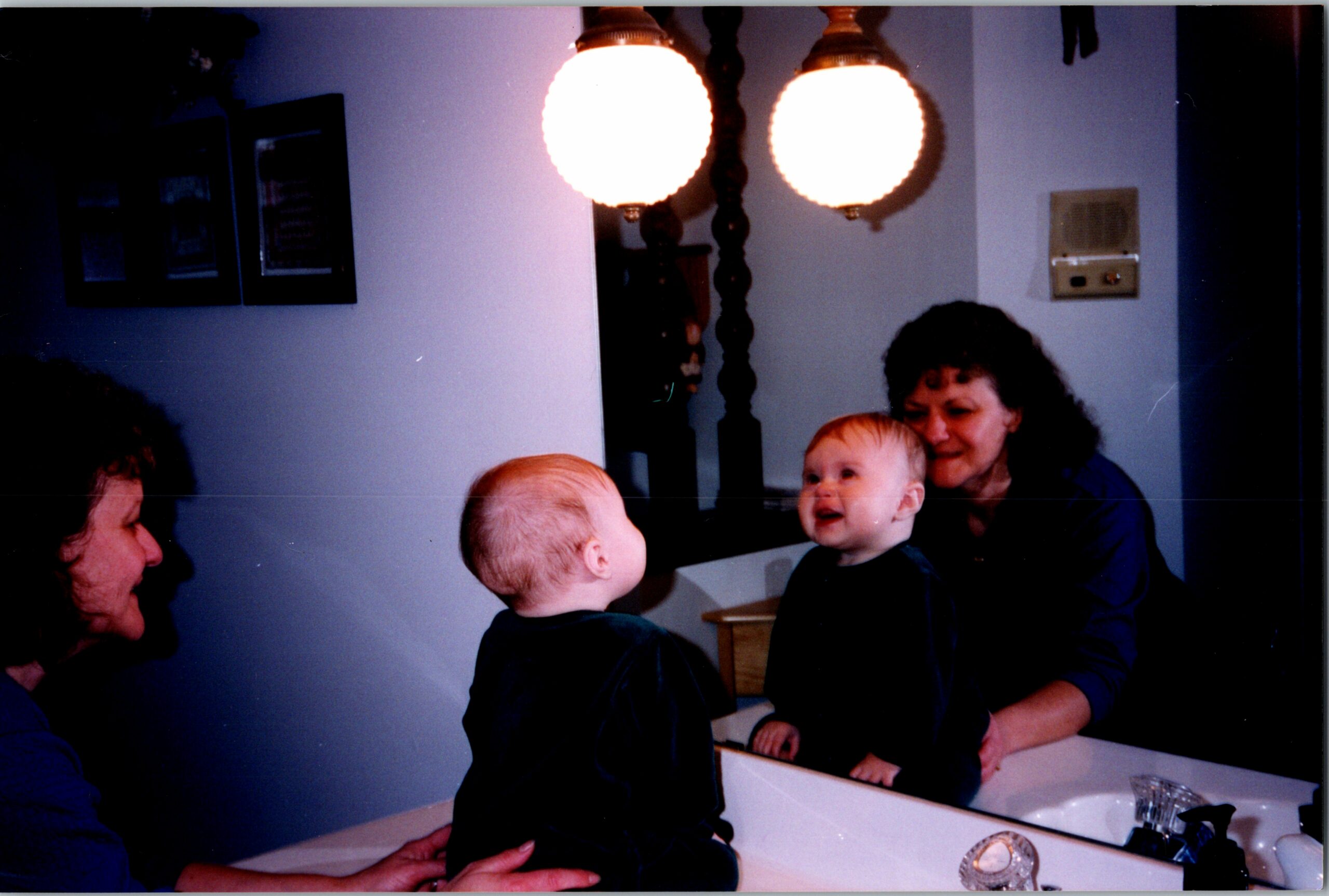 Young child sits in a bathroom sink with adults nearby, including a smiling woman in blue and another adult helping.