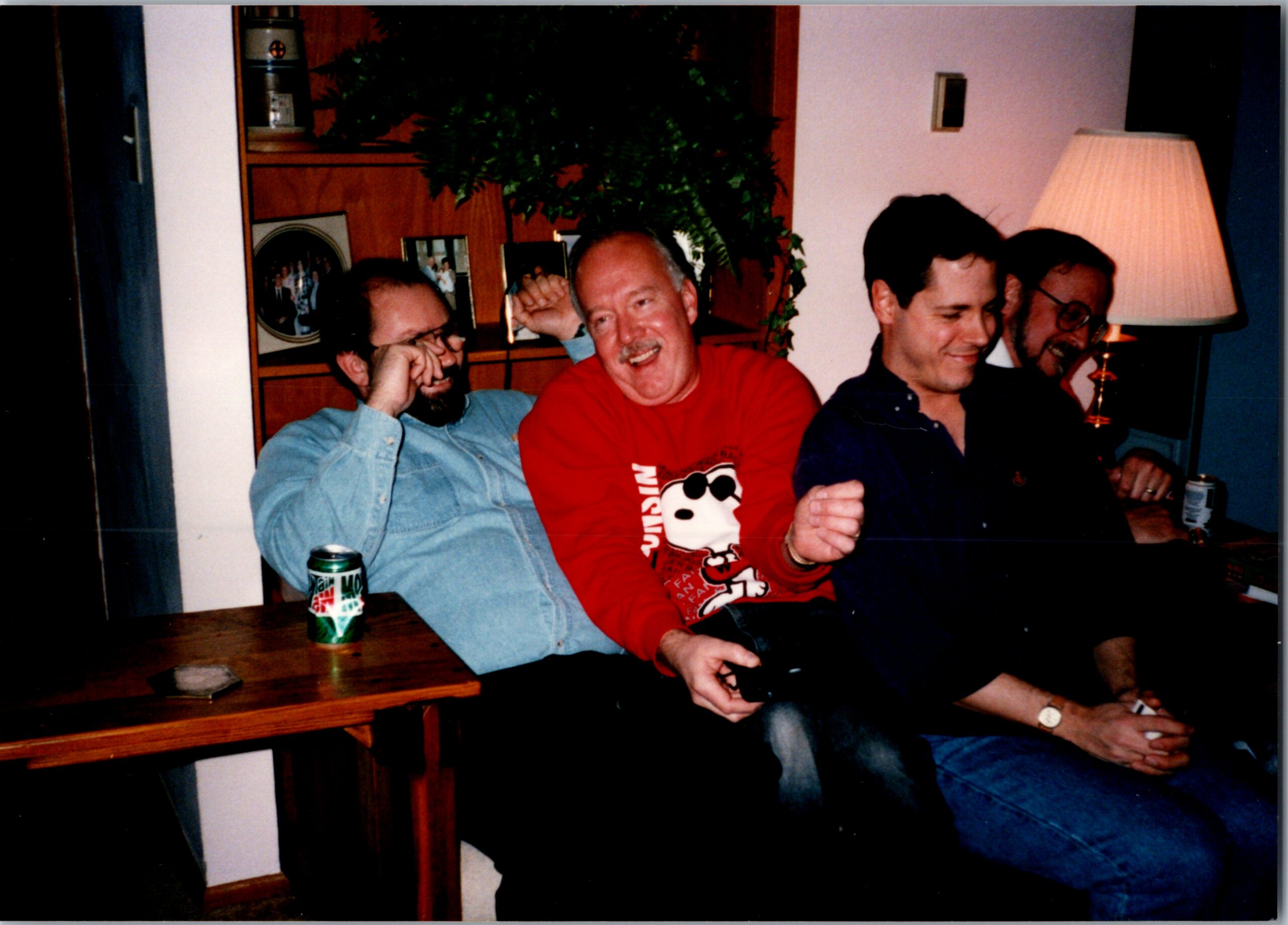 Four men sit around a table laughing together in a cozy indoor room with a lamp and plants in the background.