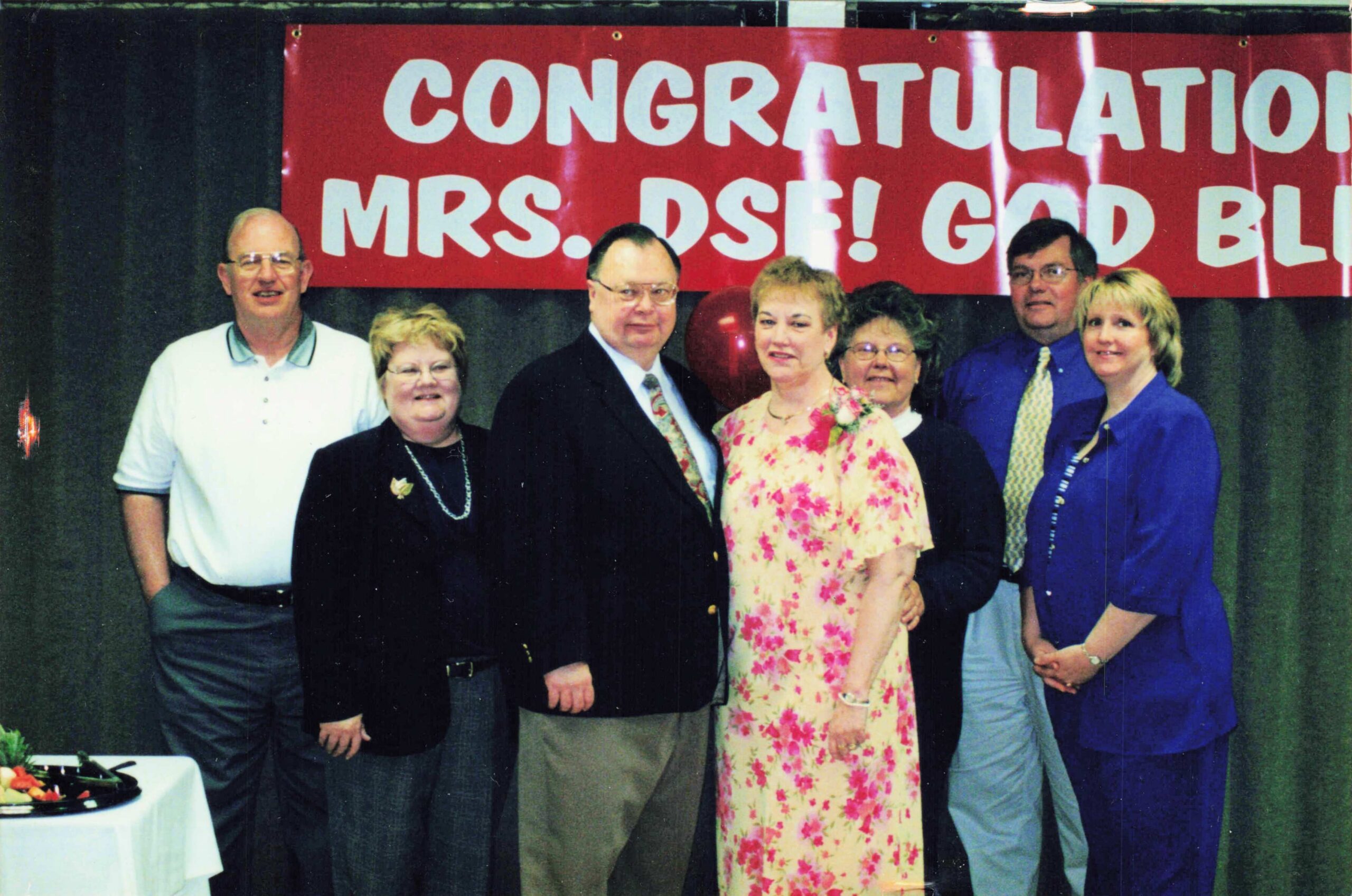 Six adults and one younger woman pose on a stage in front of a red banner that reads 'CONGRATULATIONS' in large white letters.