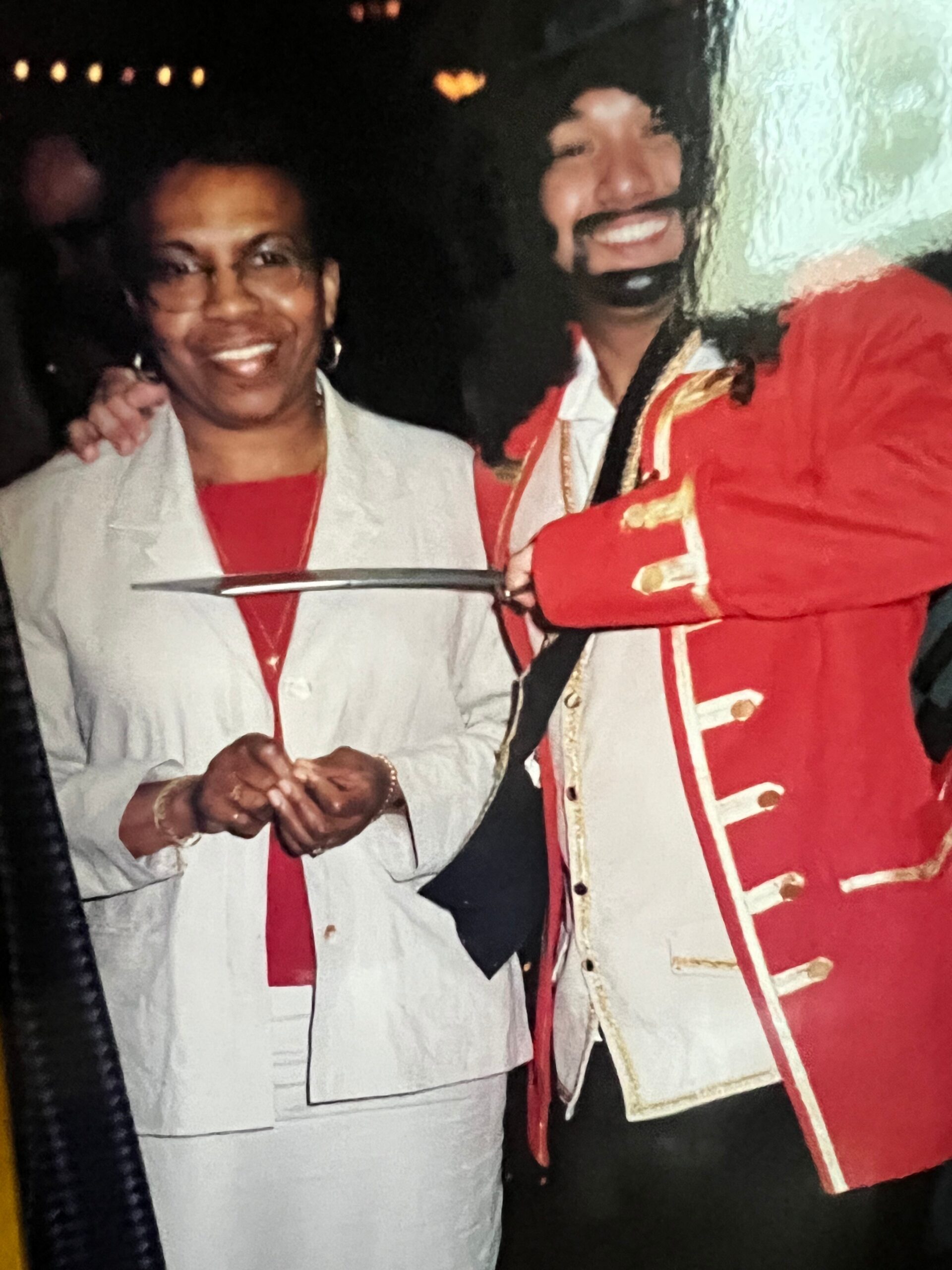 Two people smiling together at night: a person in a light gray suit with a red top and a person in a red ceremonial coat with gold trim beside them.
