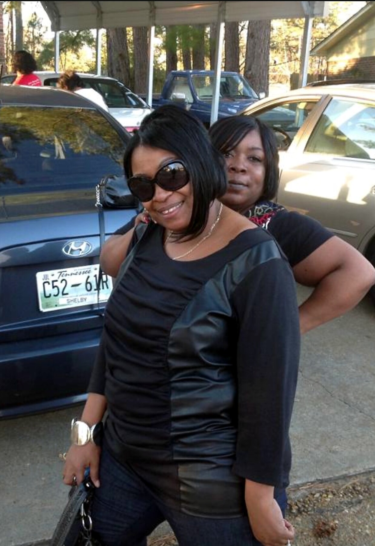 Two women pose together in a driveway next to parked cars, the woman in front wears a black dress and sunglasses, smiling at the camera.