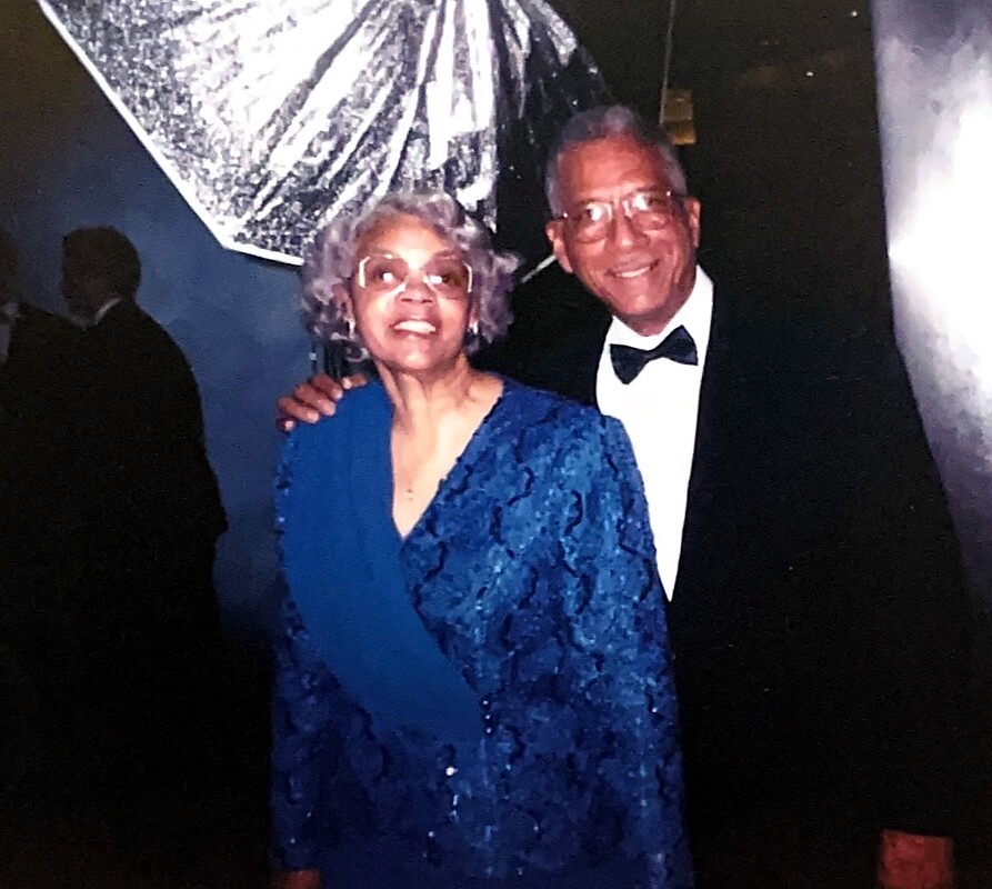 Smiling elderly couple posing together at a formal event; woman in a blue lace dress with glasses, man in a black tuxedo and bow tie, arm around her.