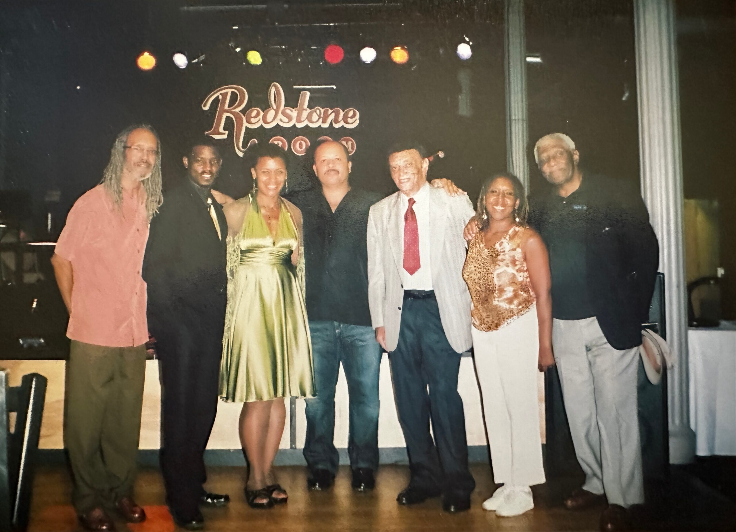 Group of eight adults posing on a stage after a performance, with colorful lights and a 'Redstone' sign in the background.