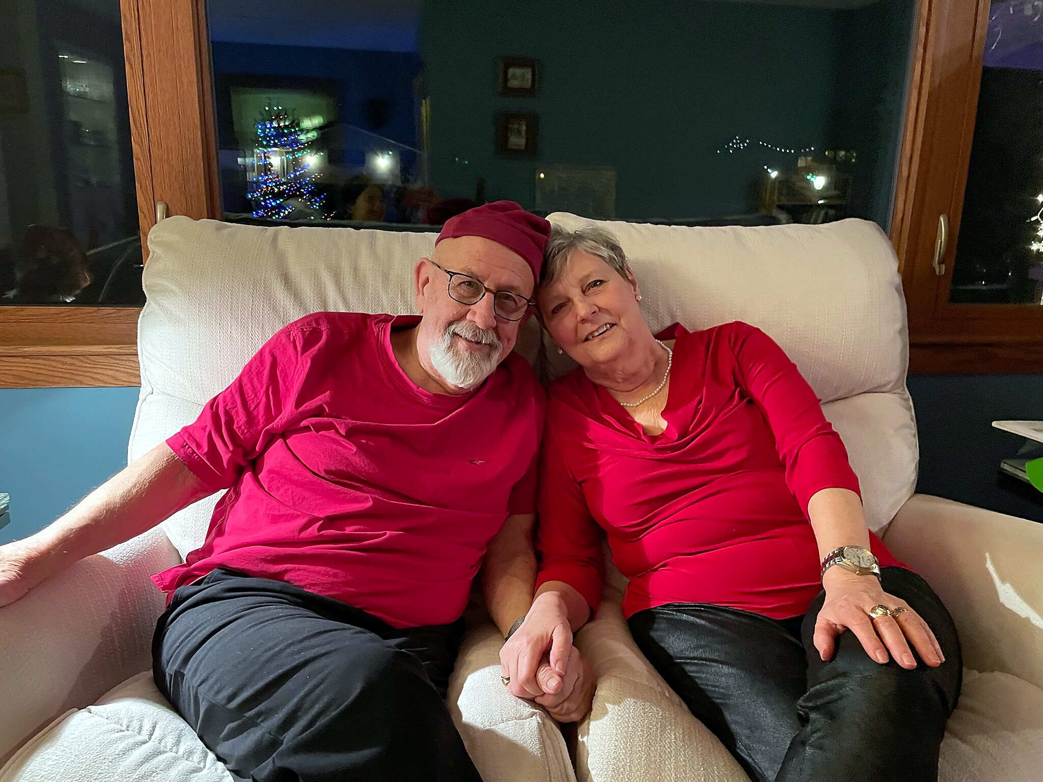 Older couple in red shirts sits close together on a beige couch, smiling at the camera with Christmas lights and tree reflected in the window behind them.”] ,