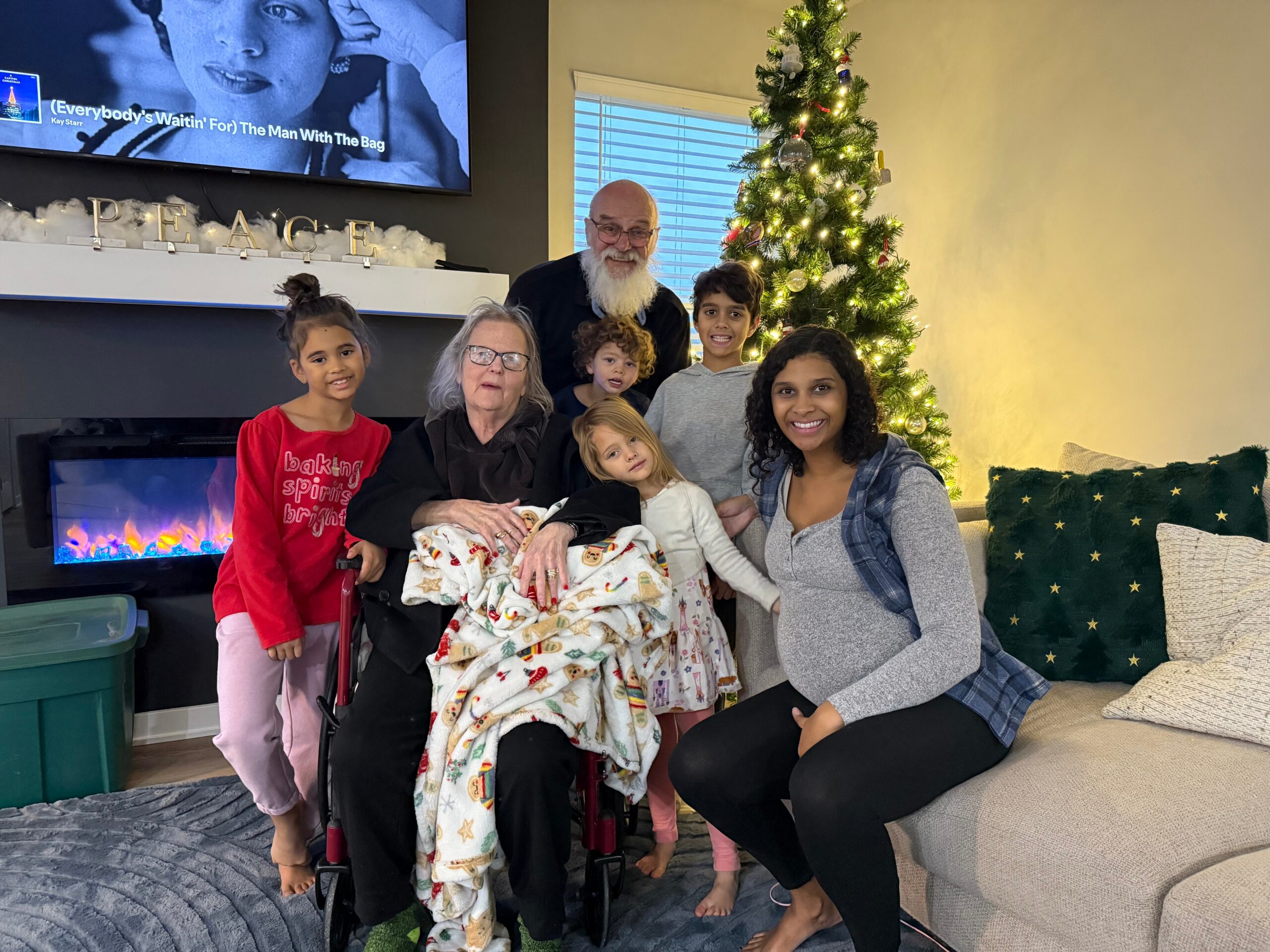 Multigenerational family posing in a living room with a Christmas tree; grandma in a wheelchair wrapped in a blanket, kids gathered around, smiling.