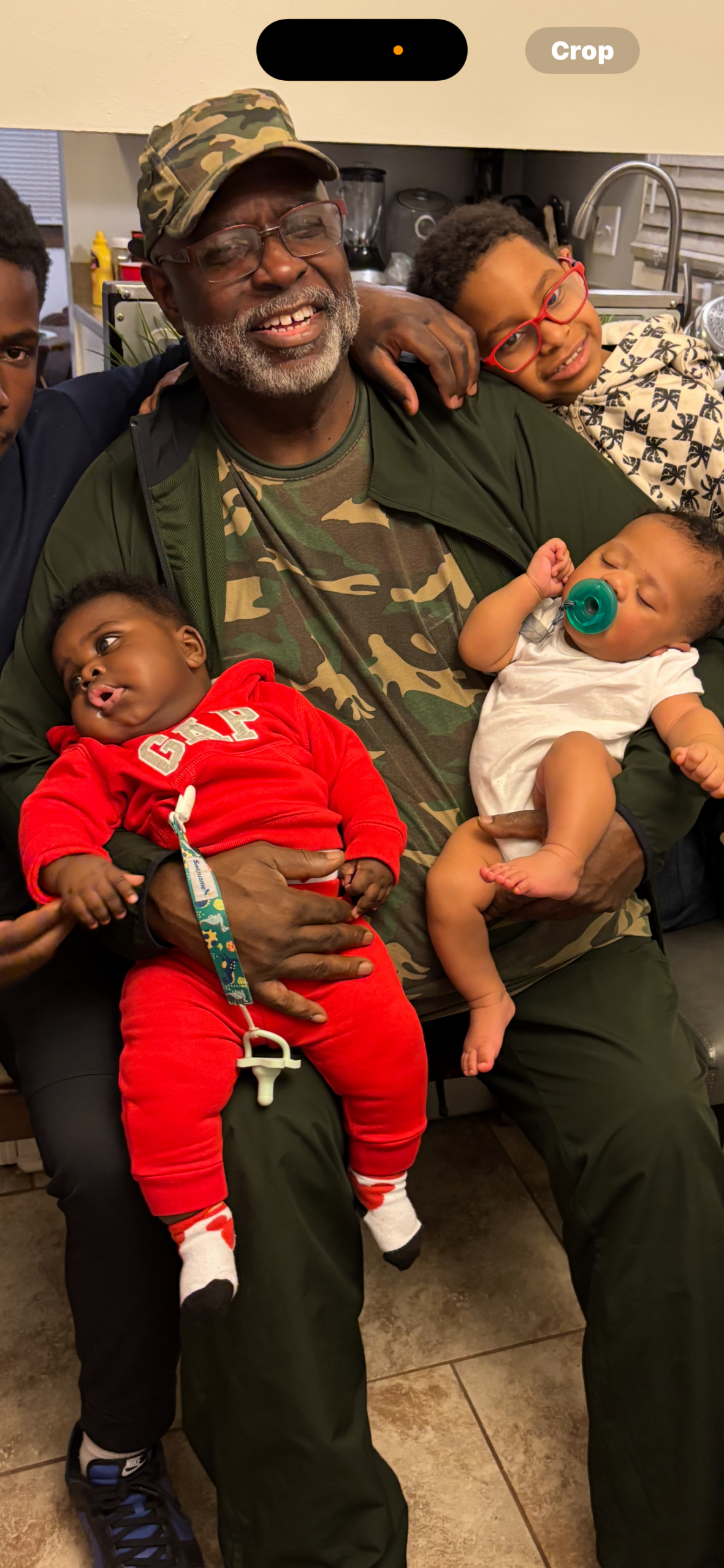 Smiling man in a camouflage cap holds two babies: a baby in red with a pacifier clip on his lap and a baby in a white onesie with a green pacifier, in a kitchen.
