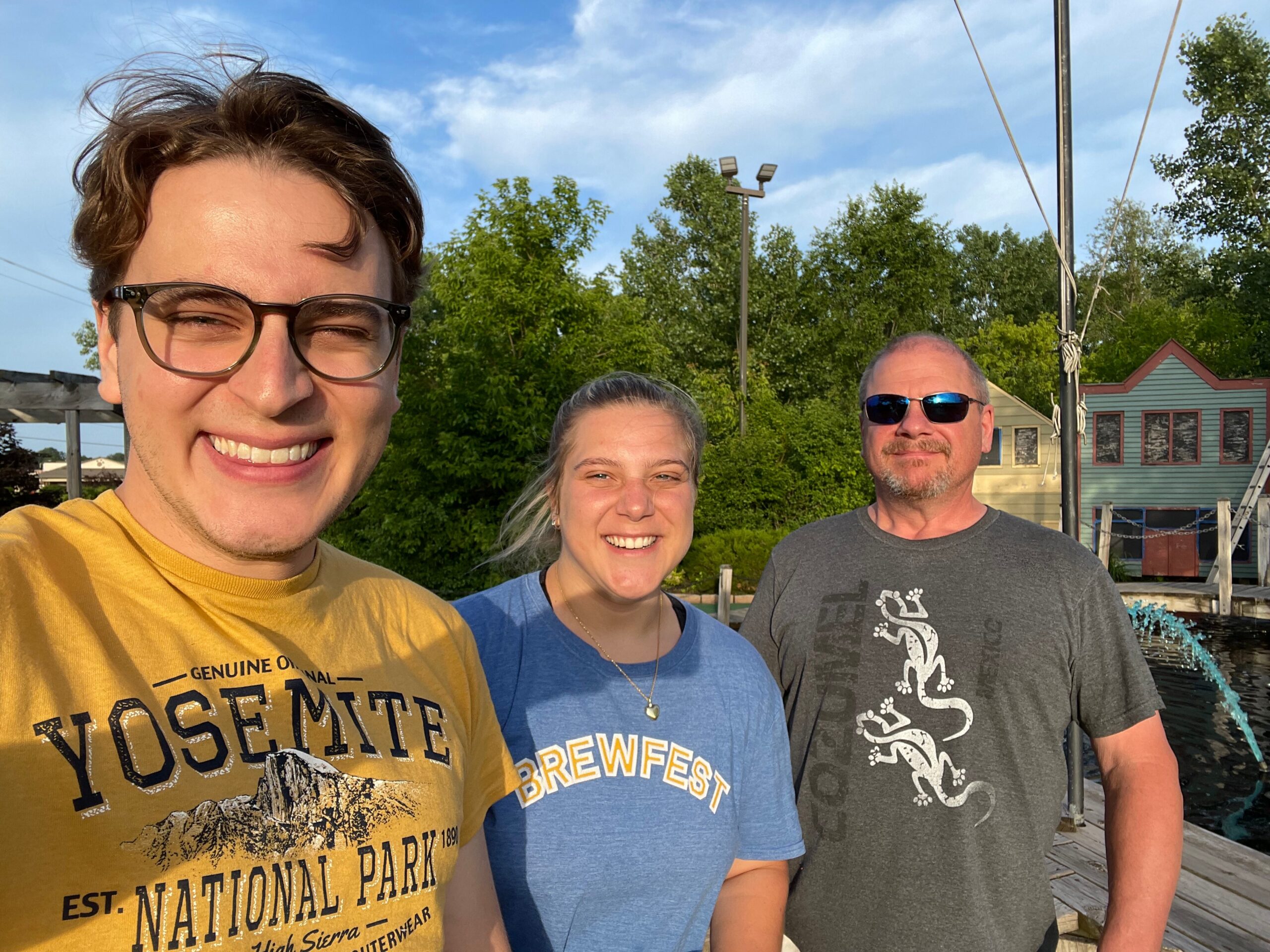 Three adults posing for a sunny outdoor selfie: left person in a yellow Yosemite National Park shirt, middle person in a blue Brewfest shirt, right person in a gray dragon-themed shirt, with trees and a small house in the background.