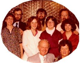 Group portrait of ten adults in a warmly lit room, smiling at the camera, arranged in two rows by the circular frame.