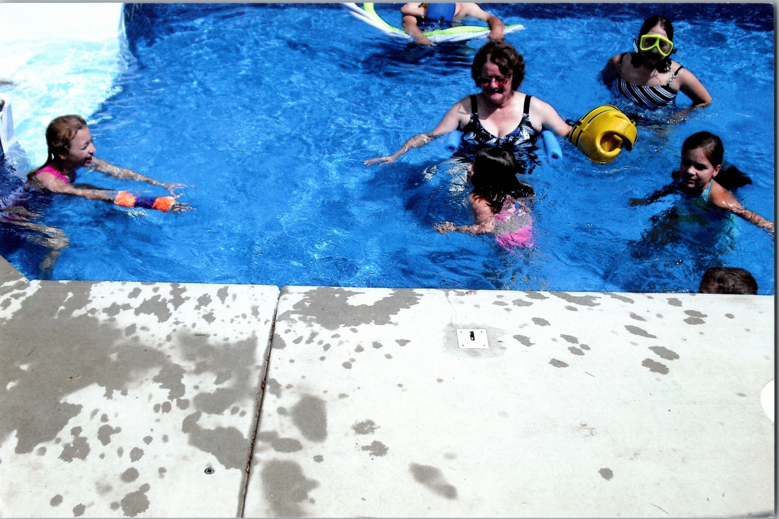 Group of people swimming in a bright blue pool, including a woman with blue arm floaties and children nearby with swim gear.