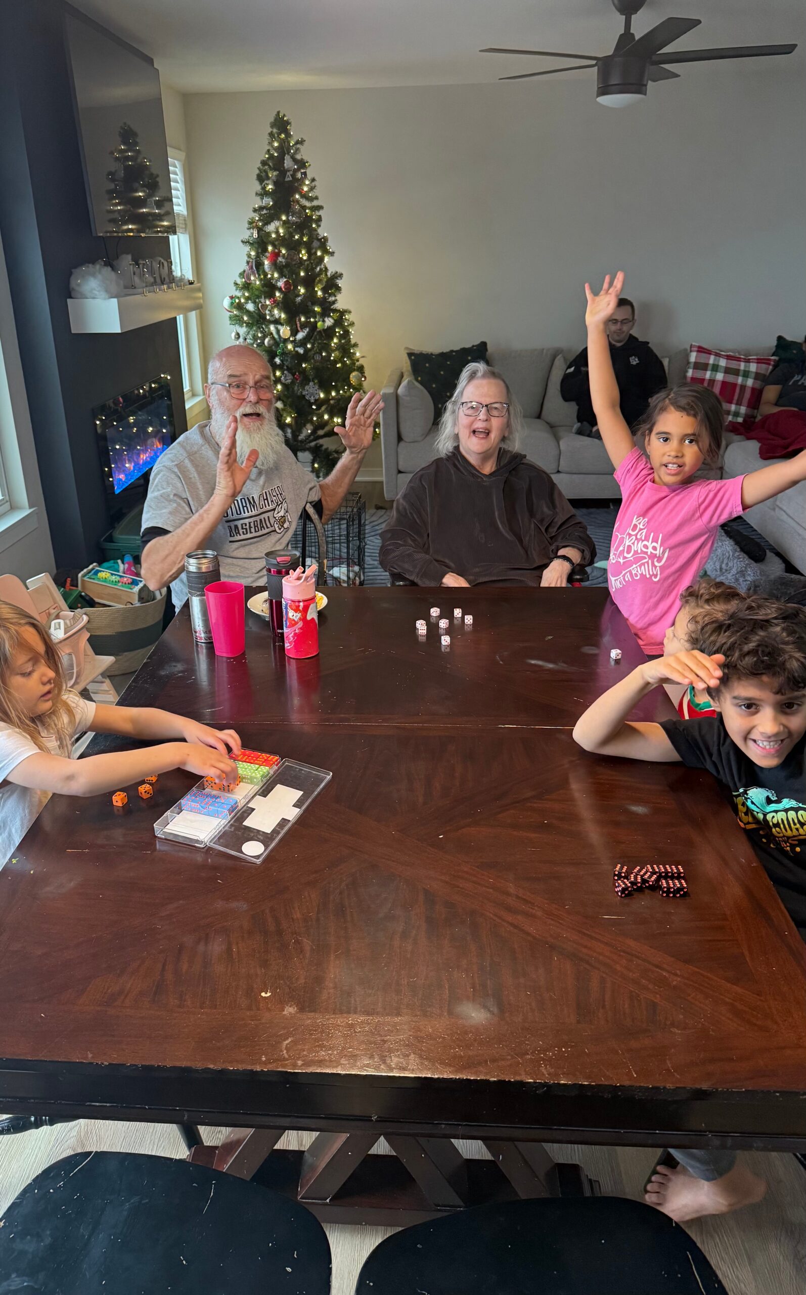 Family at a dining table playing dice games indoors, Christmas tree in the background.