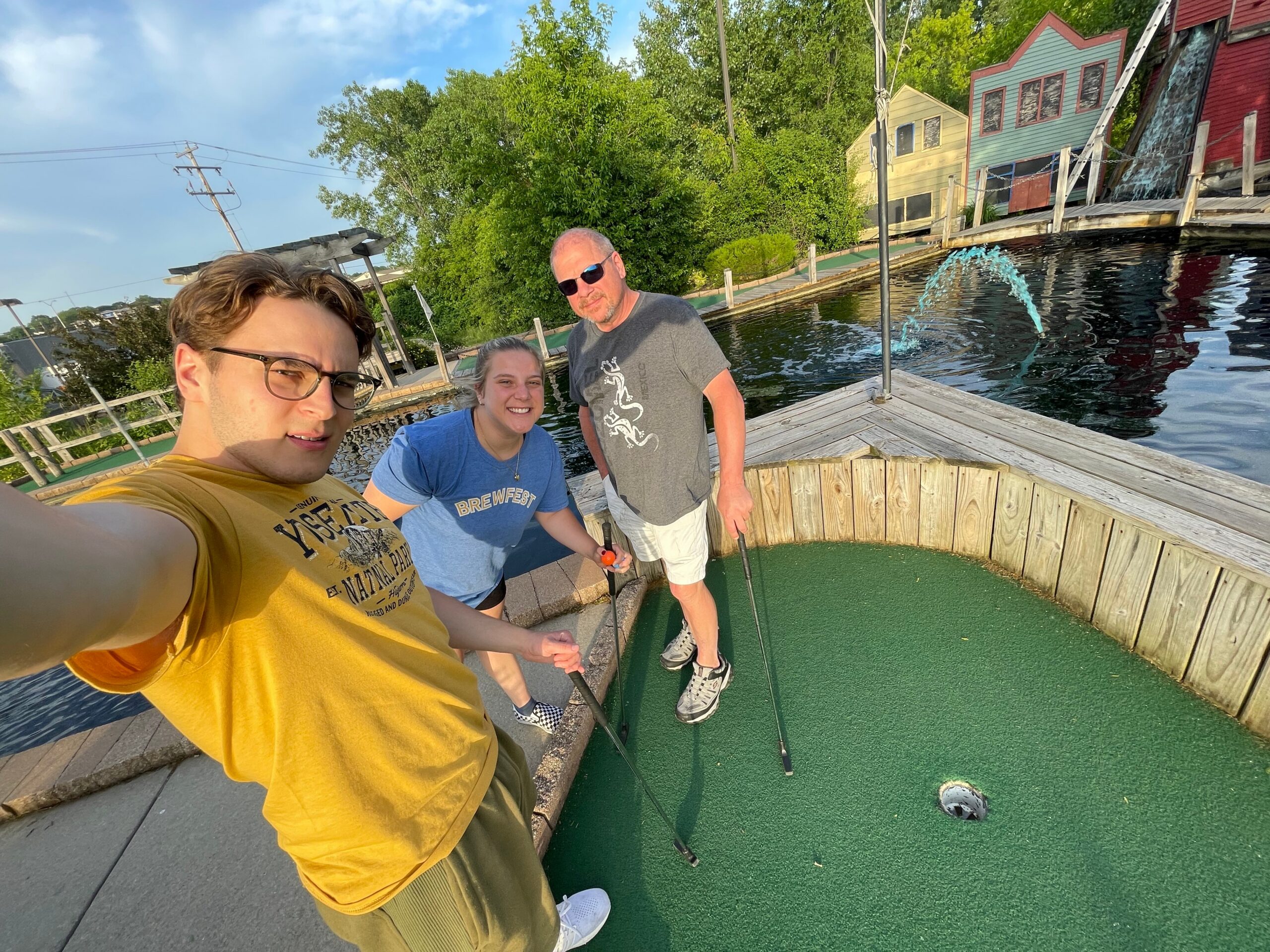 Three friends taking a selfie at a mini golf course with green turf, a small pond, and colorful buildings in the background.