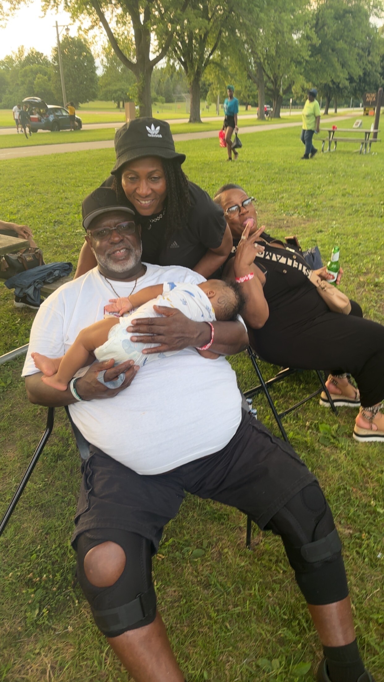 Smiling man in a white shirt holds a baby as two women lean in close in a sunny park setting with green grass and trees behind them.