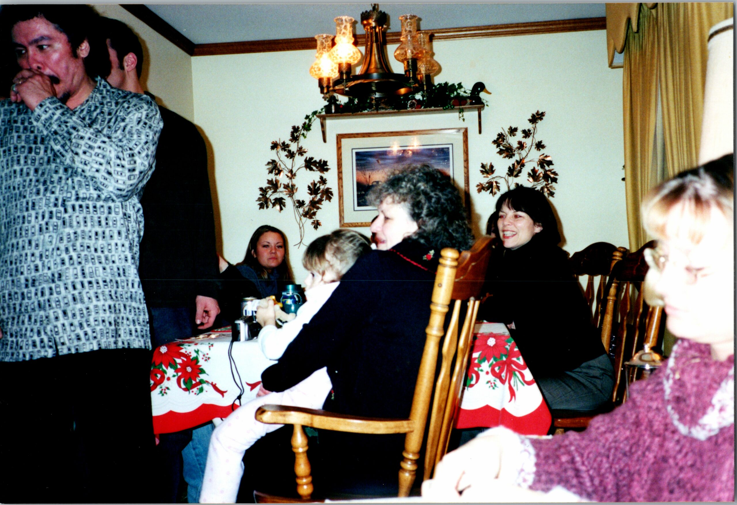 People seated around a Christmas dinner table, with a festive red‑green tablecloth; a man in a patterned shirt stands on the left.