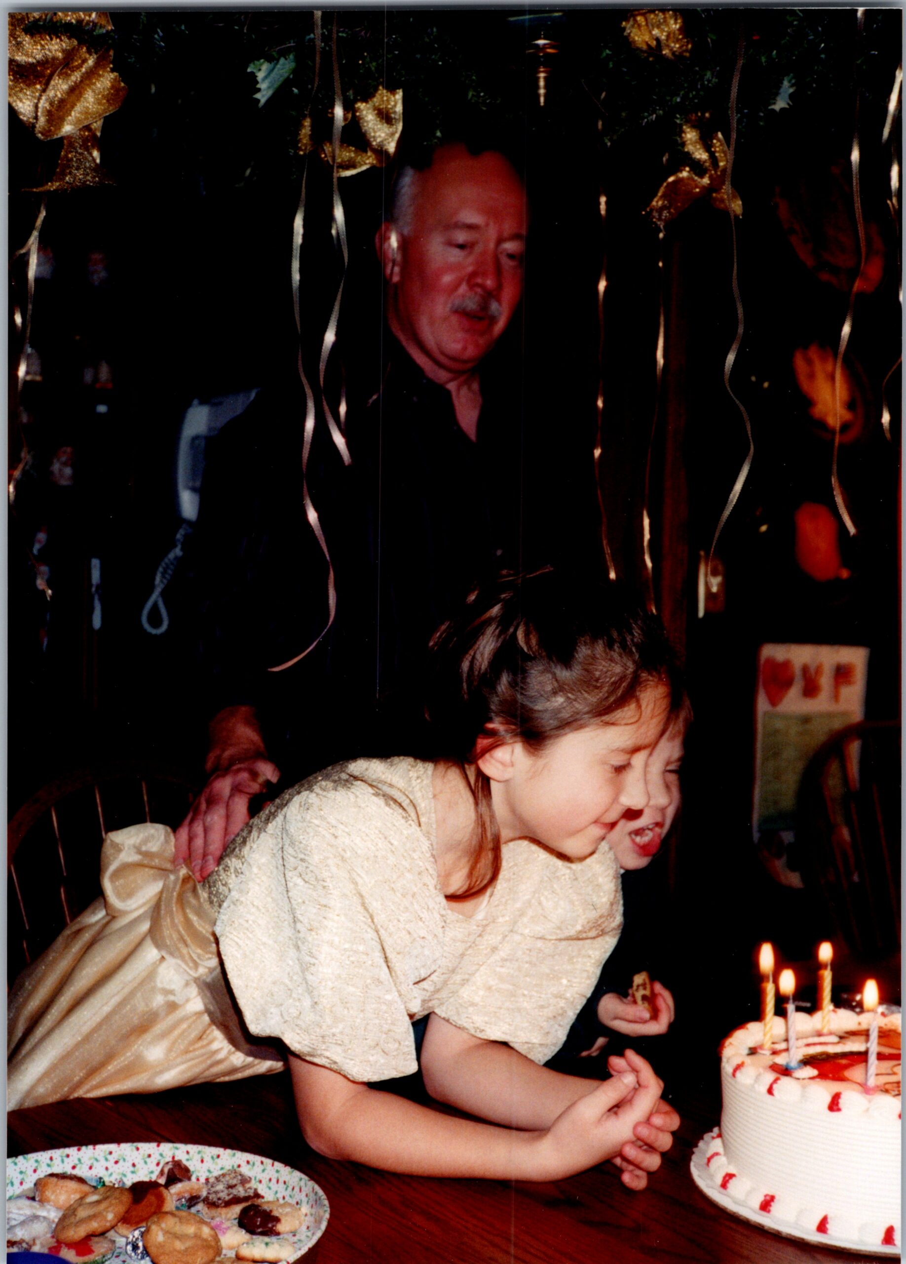 Young girl in a cream dress leans toward a birthday cake with lit candles as an older man looks on in the background, celebrating.