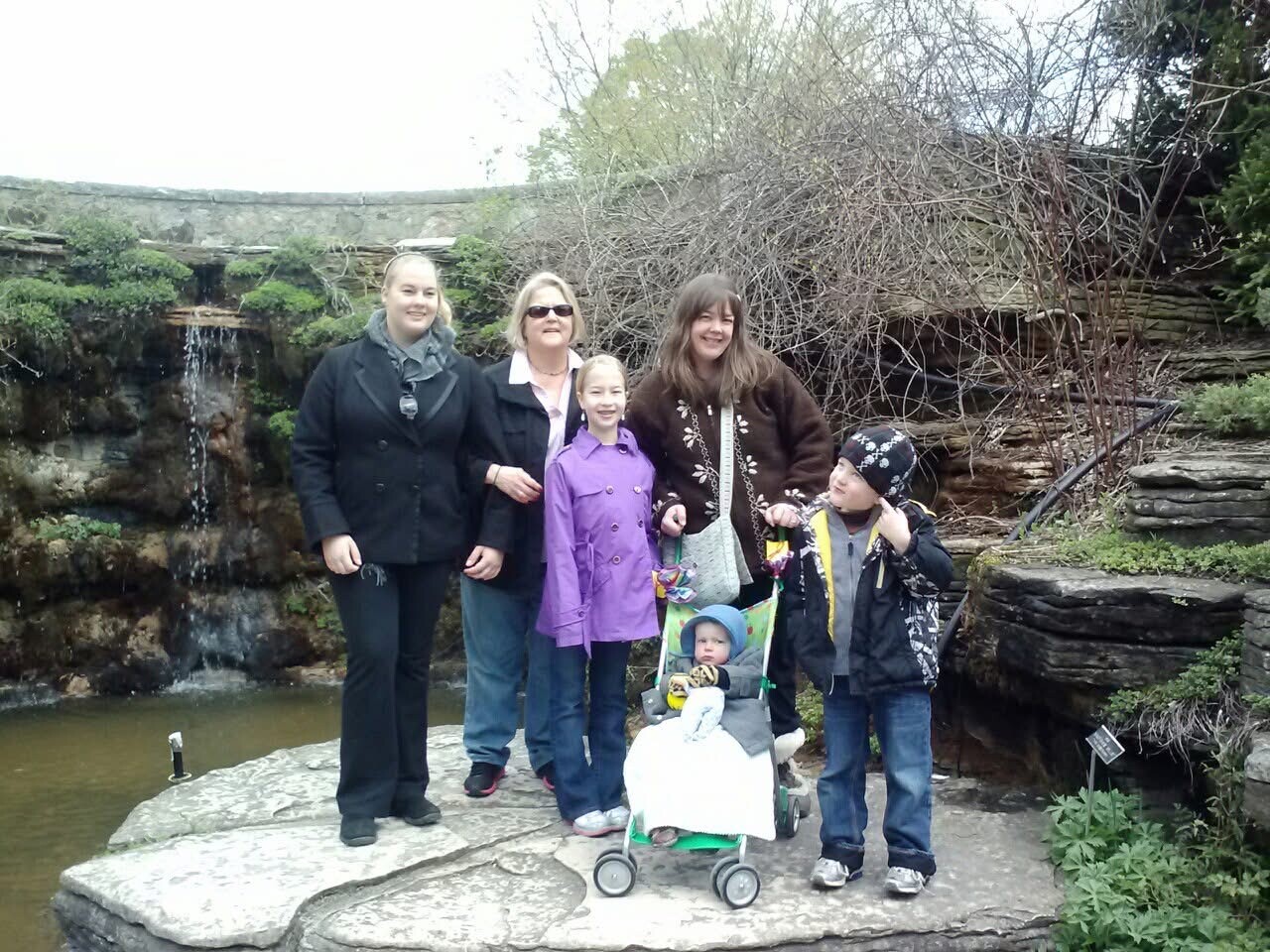 Family group posing on a stone patio by a small waterfall and rock wall in a garden; a baby in a stroller sits at the front.