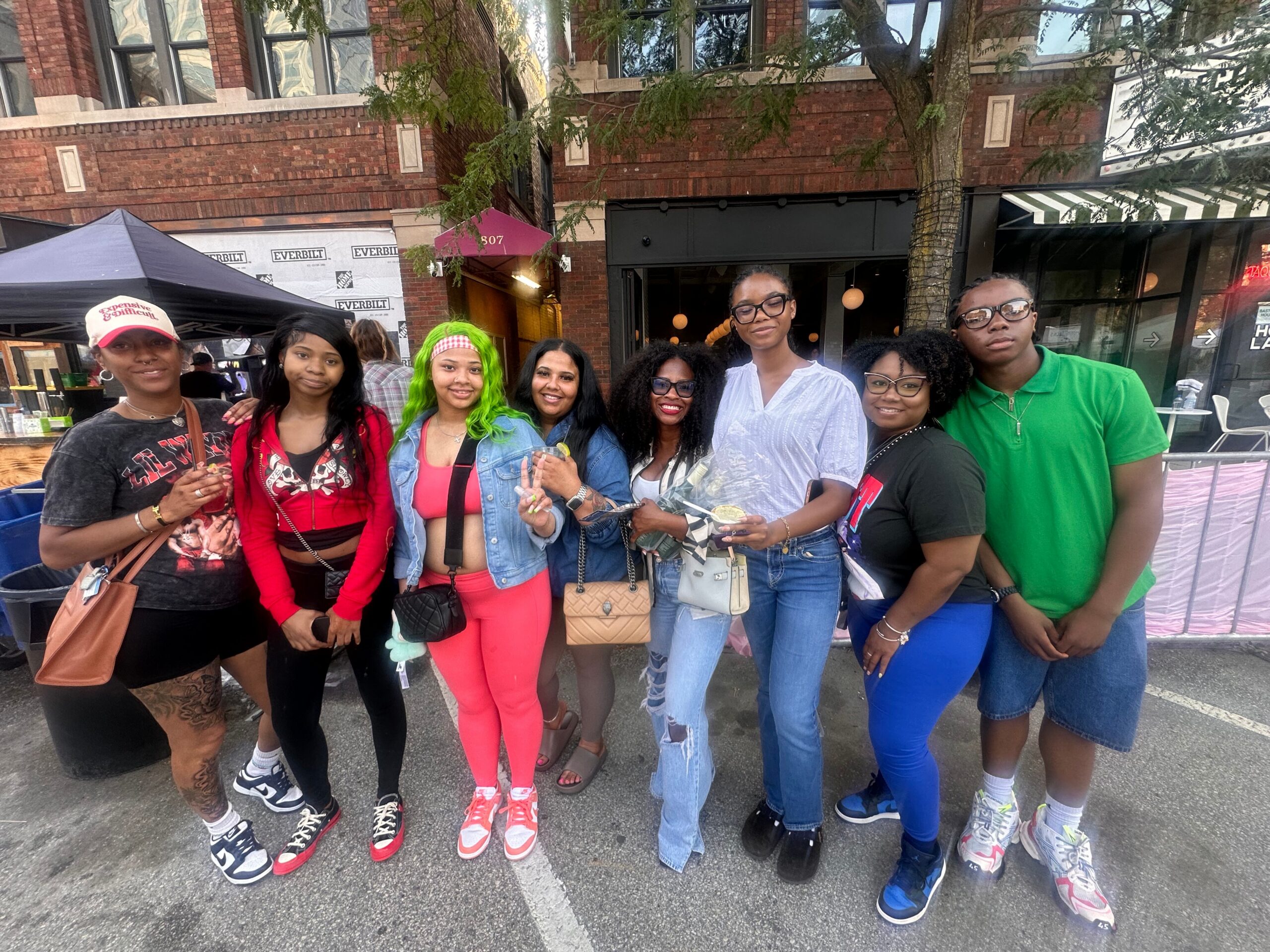 Group of eight friends posing together on a city street at an outdoor festival, brick buildings in the background.