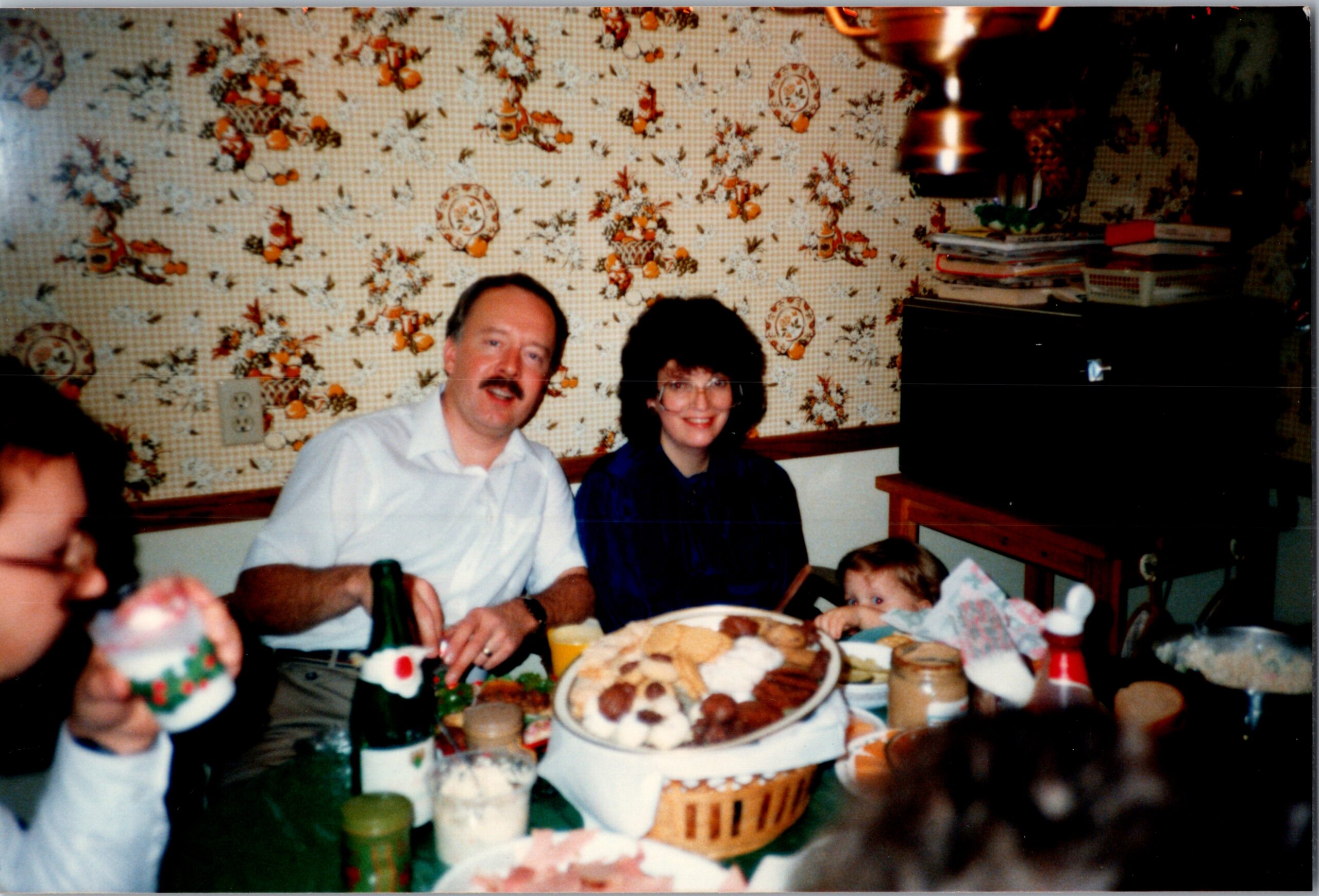 Family gathered around a dining table for a celebration with cake, drinks, and snacks; man with mustache and woman smile at the camera, a child nearby, pastel floral wallpaper in the background.