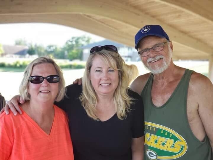 Three adults posing together, two women and one man, smiling under a wooden canopy near the water.