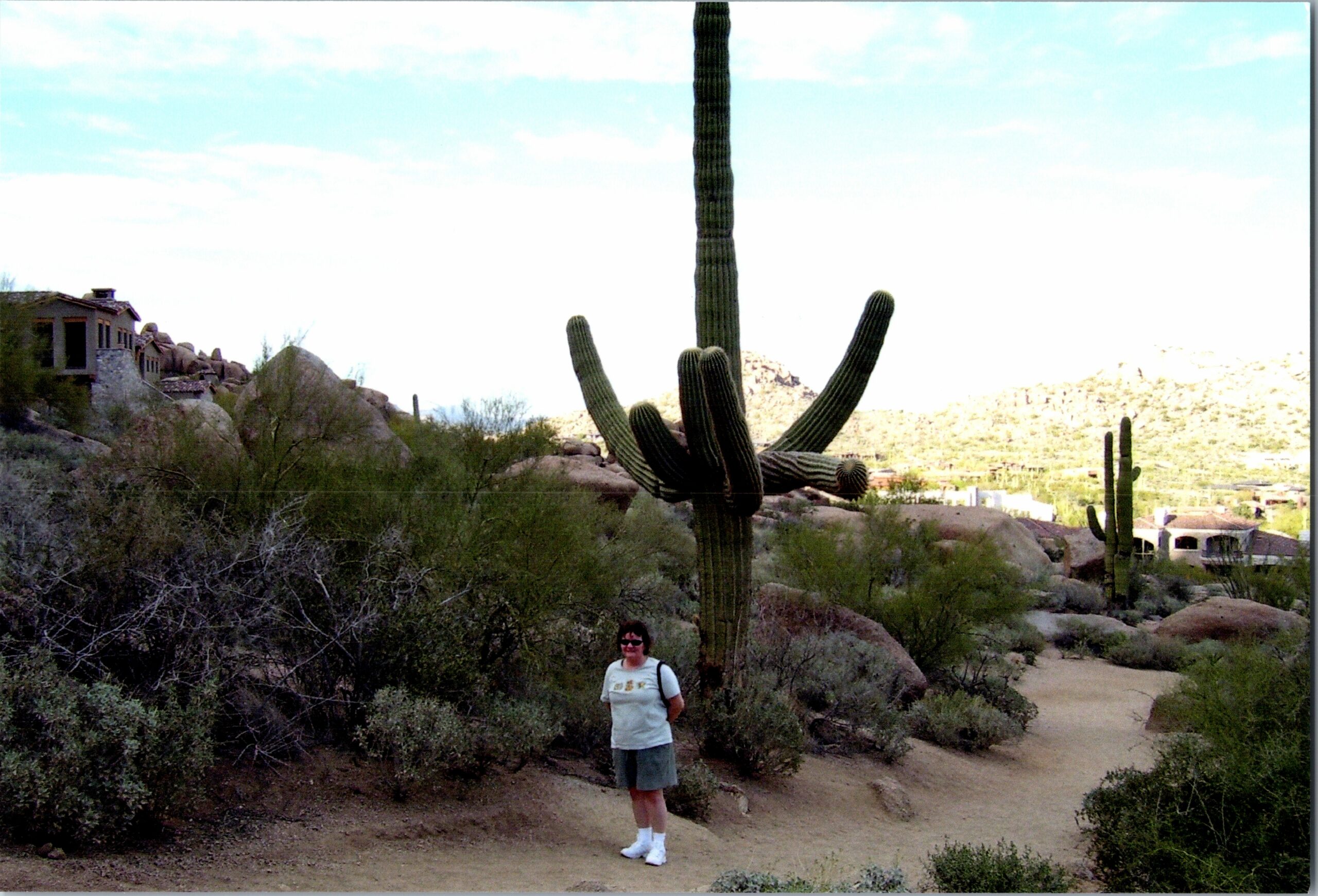 Woman standing on a desert trail beside a tall cactus, with rocky hills and houses in the distance