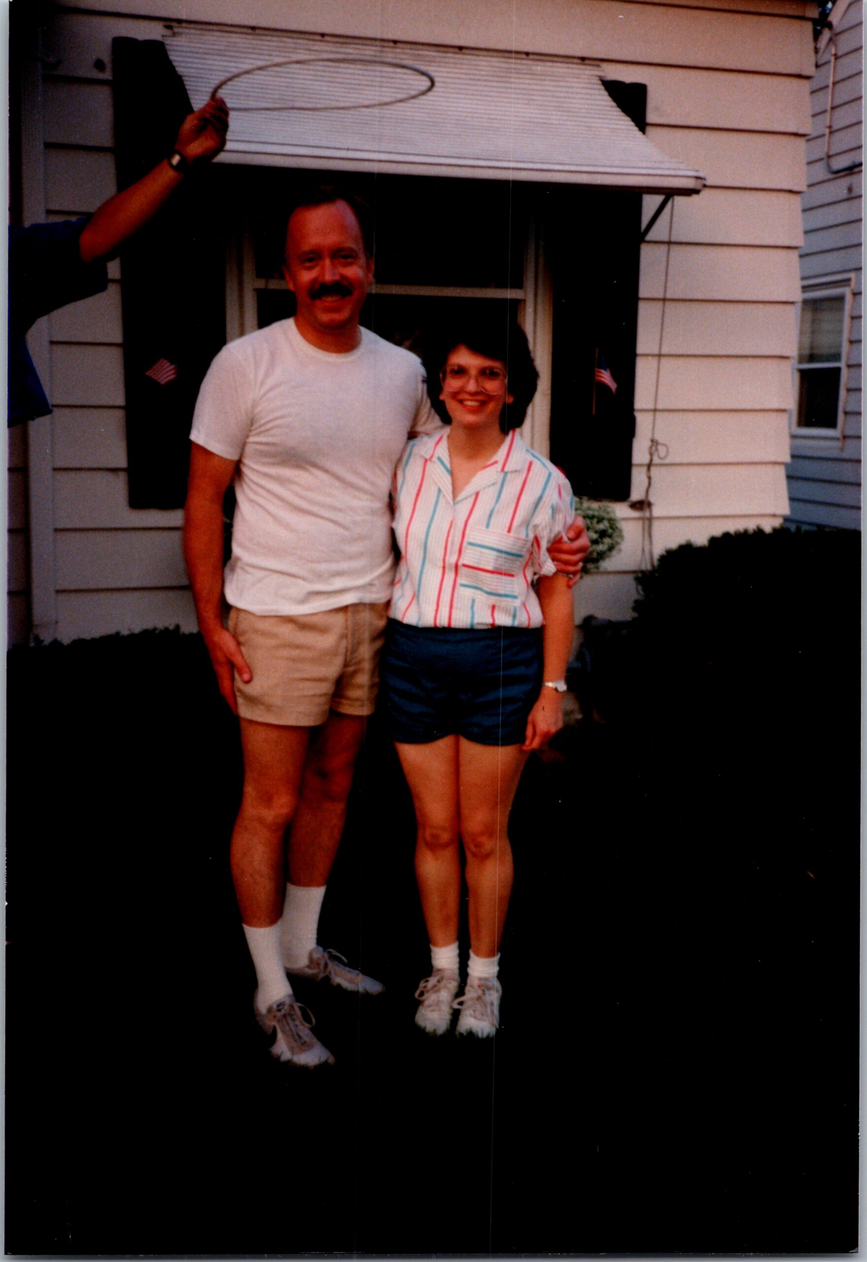 Two adults pose together outside a house, smiling at the camera in casual summer clothes (t-shirt/shorts for him, striped blouse/shorts for her). A third person’s hand holds a hula hoop overhead.