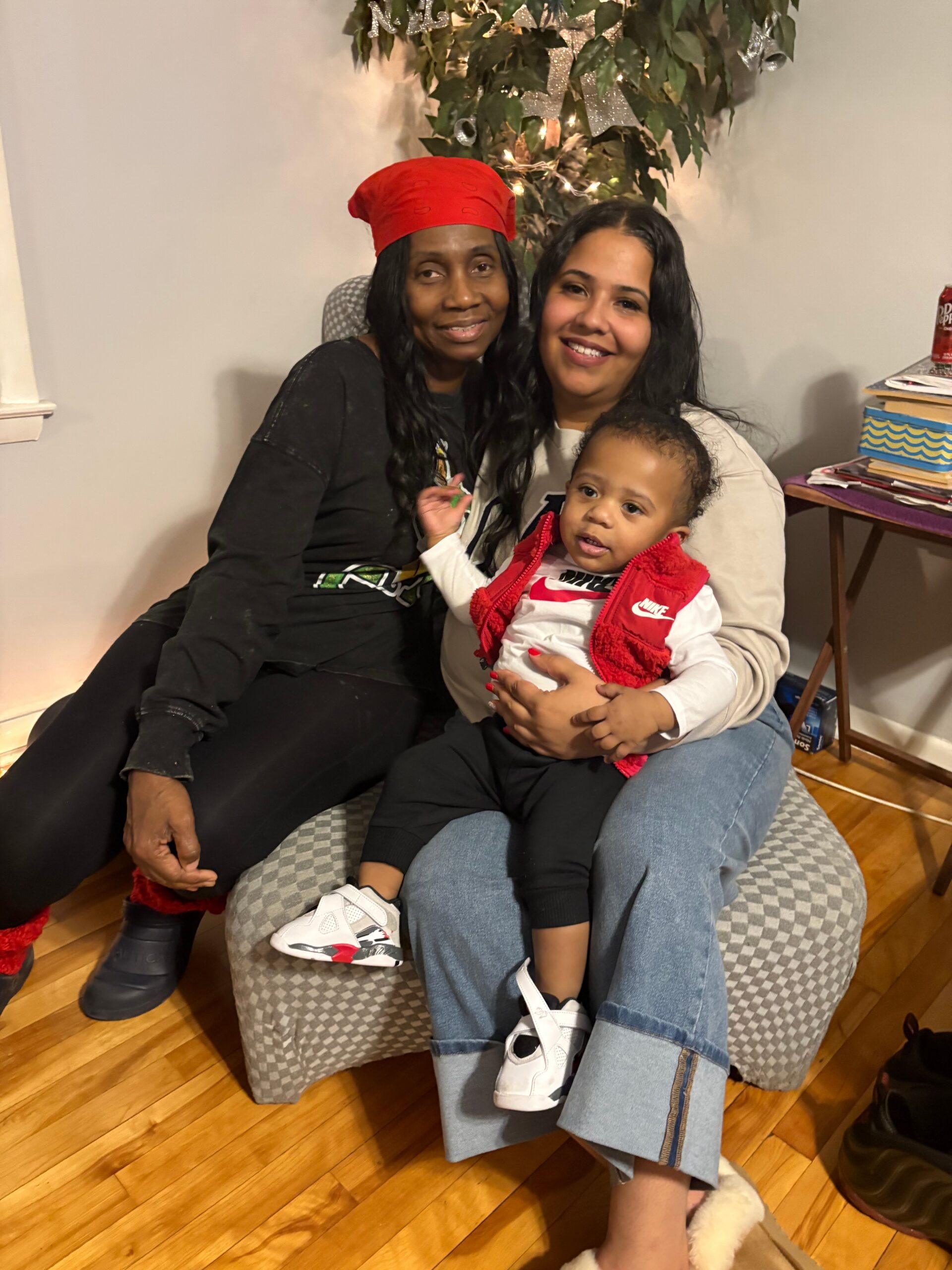Three people seated together on a checkered ottoman, smiling for a casual family photo indoors with holiday decor in the background; a child sits on a woman's lap wearing a red vest.