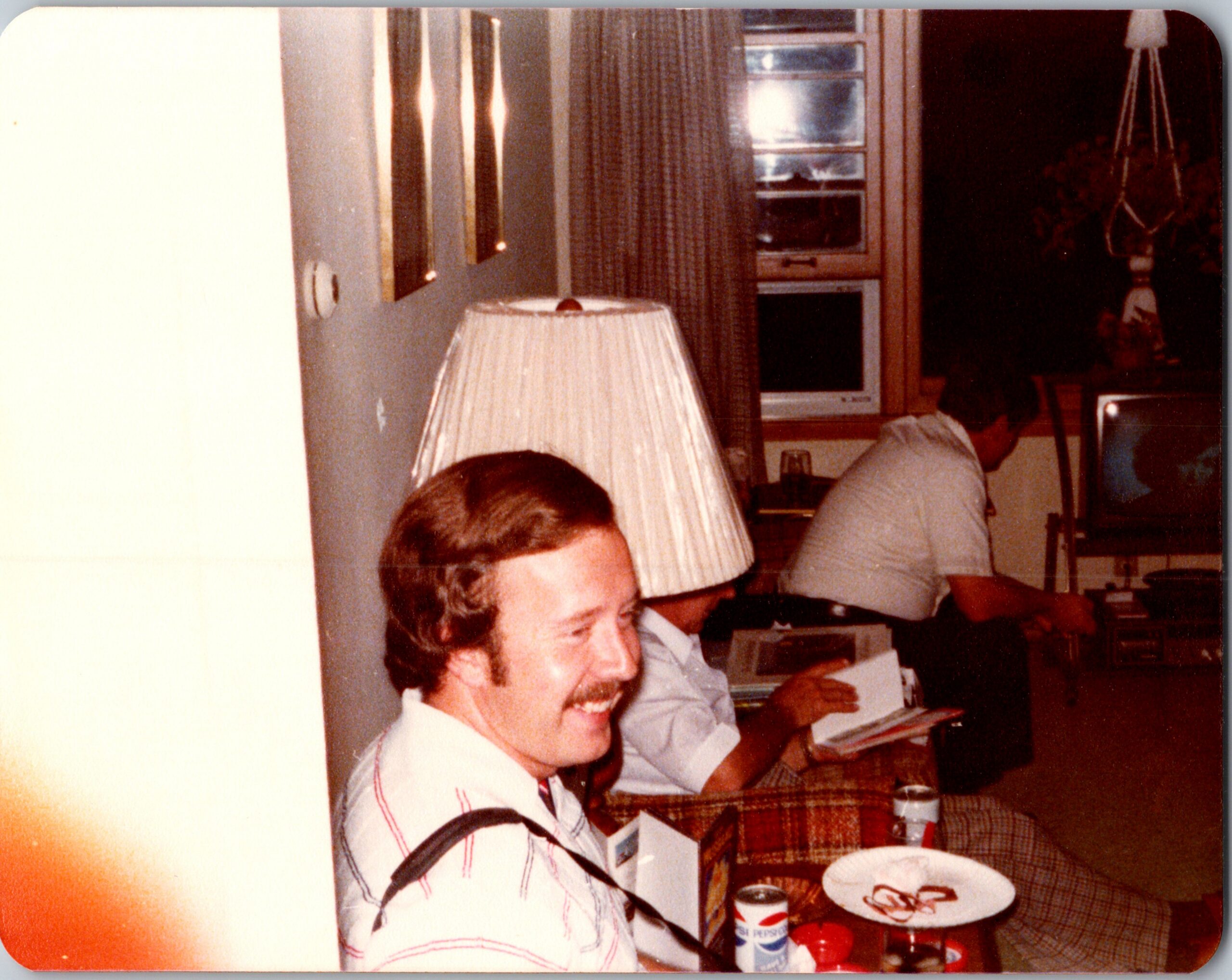 Man with a mustache smiling at a dining table with plates and Pepsi cans in a retro kitchen/dining area.