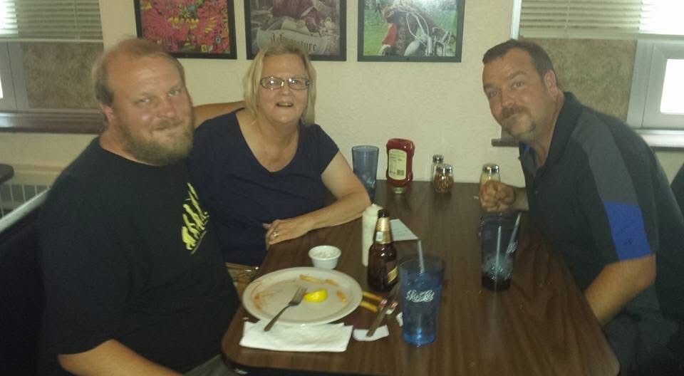 Three adults sit around a casual restaurant table, smiling at the camera; plate with leftover food and utensils in the foreground, plus bottles and cups on the table.